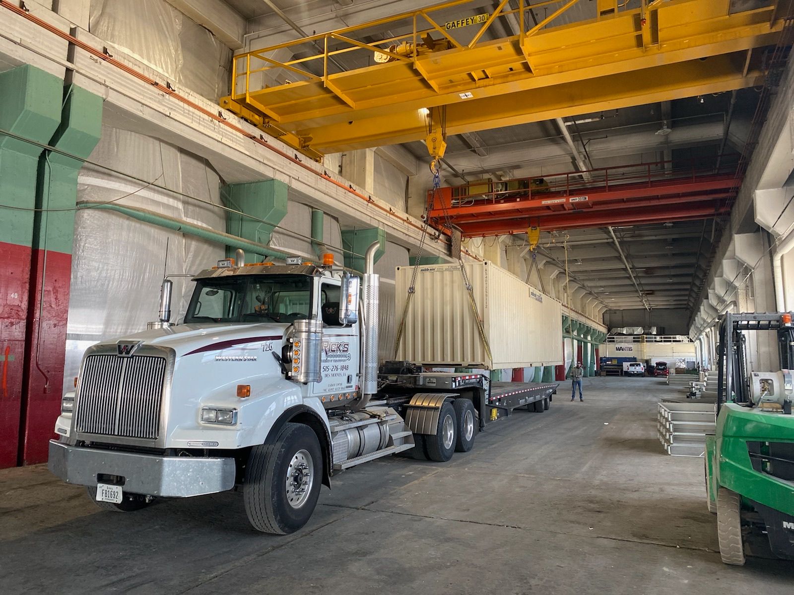 A semi truck is parked in a warehouse under a crane.