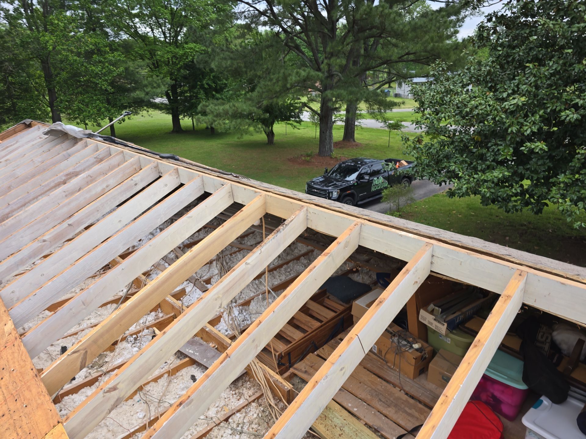 Roof structure under construction, with exposed beams and insulation, overlooking a yard and trees.