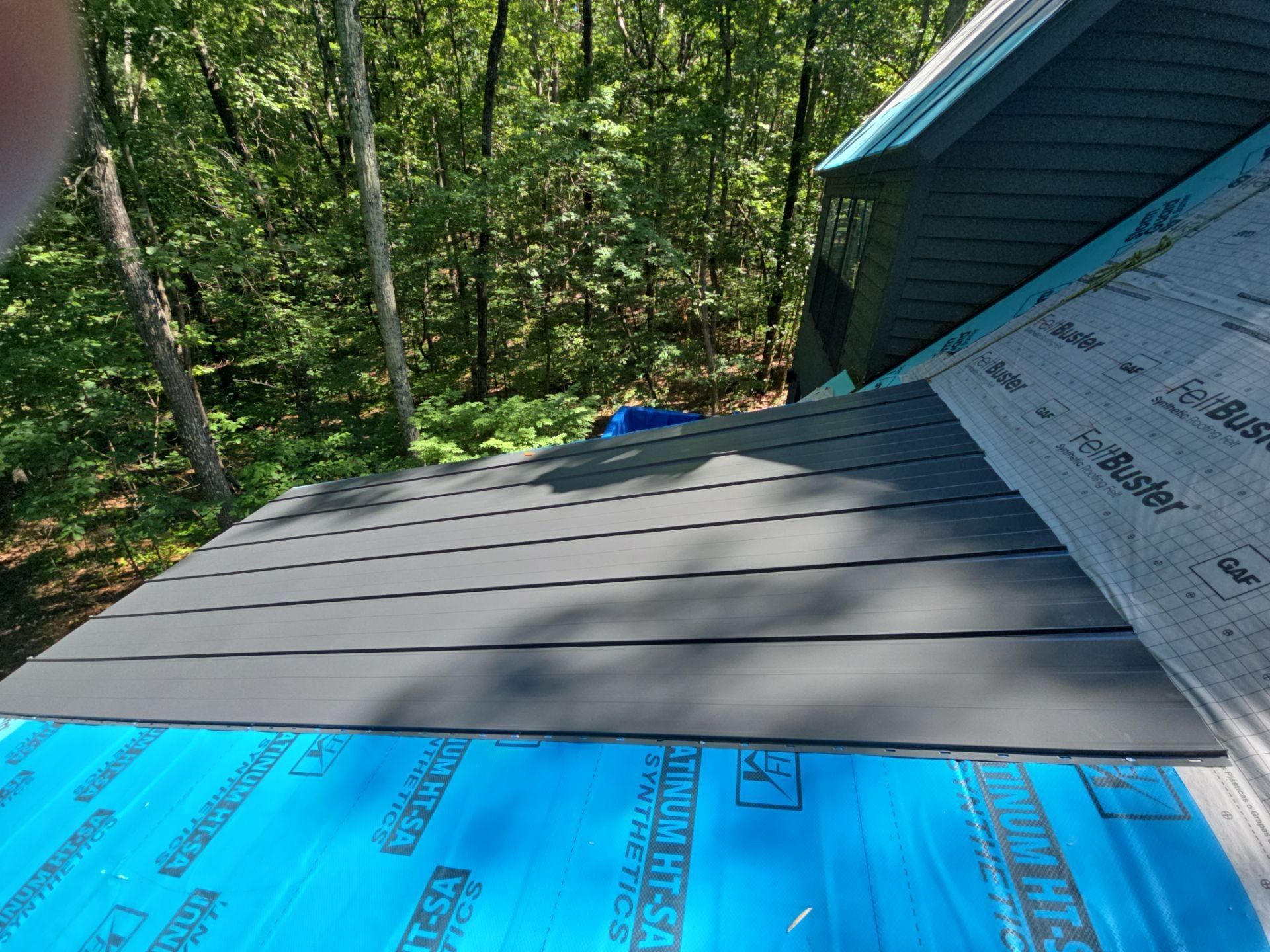 Roofing work in progress, dark gray panels over blue waterproof underlayment, set in a wooded area.