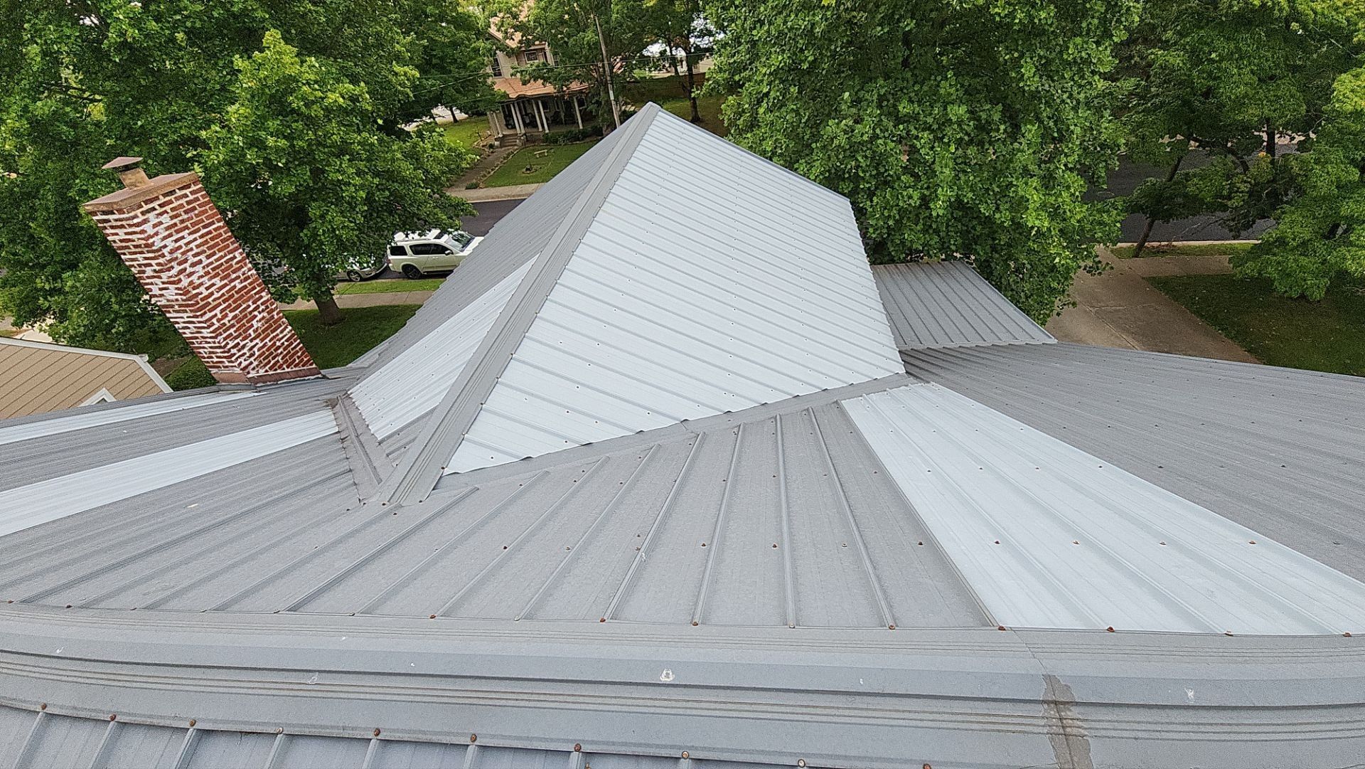 Gray metal roof with a central pyramid-shaped section surrounded by trees.