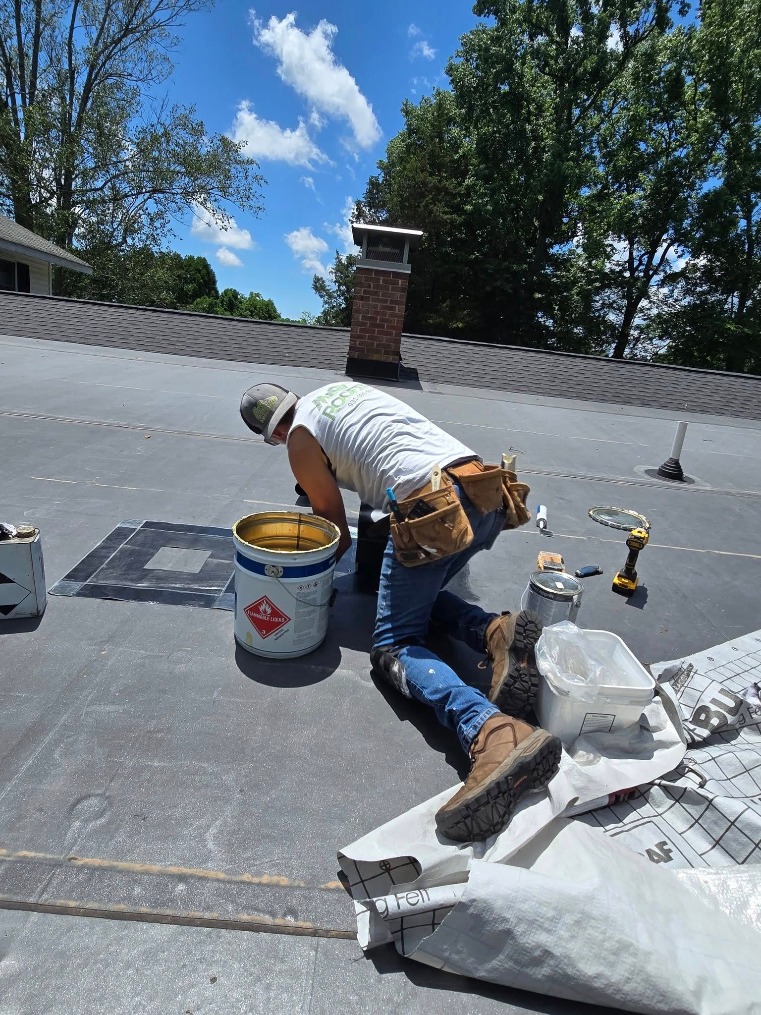 Roofer on a flat roof, working near a chimney, with tools and materials on a sunny day.