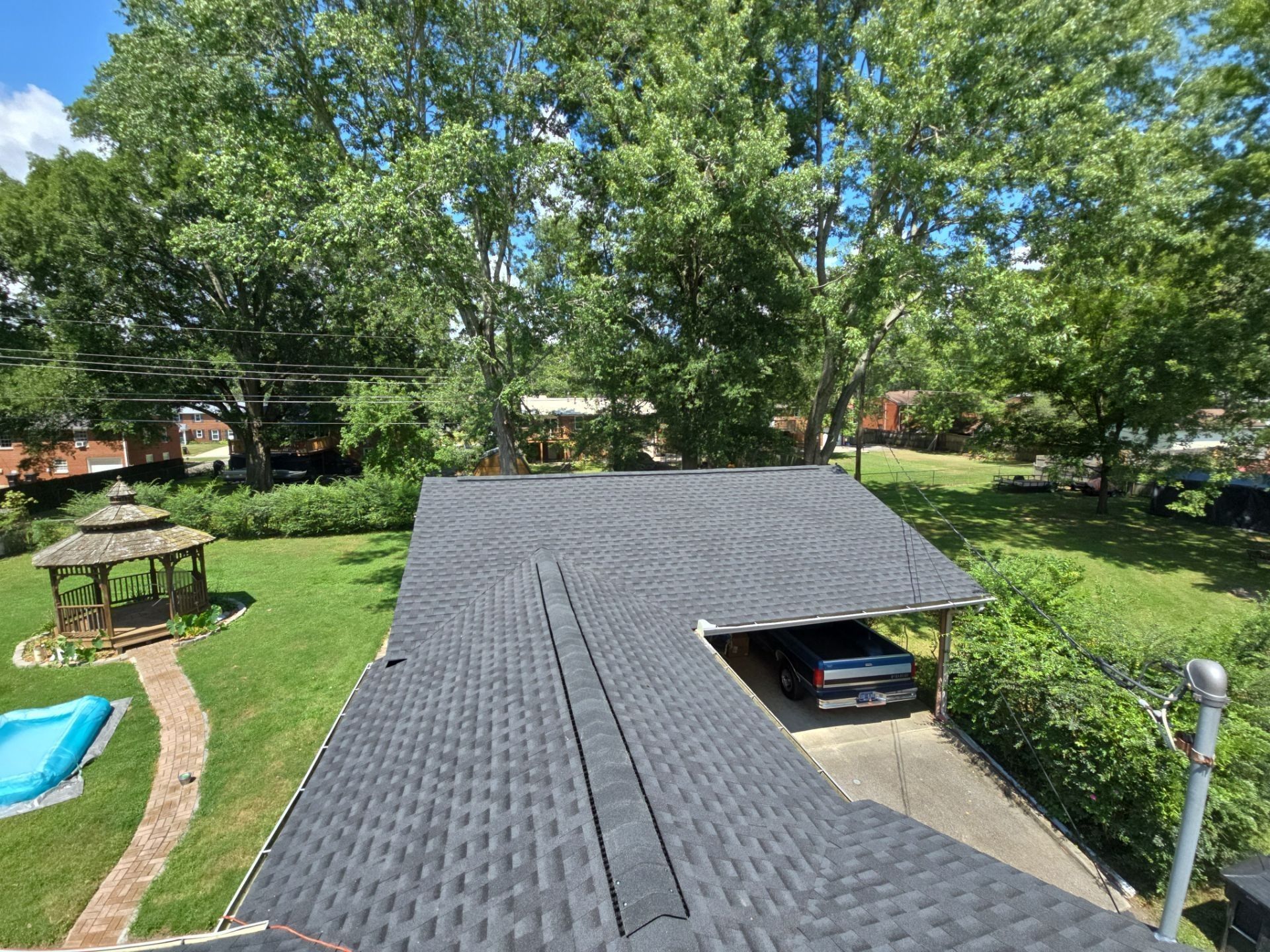 View from roof of house with carport. Dark gray shingles. Green lawn, trees, gazebo.