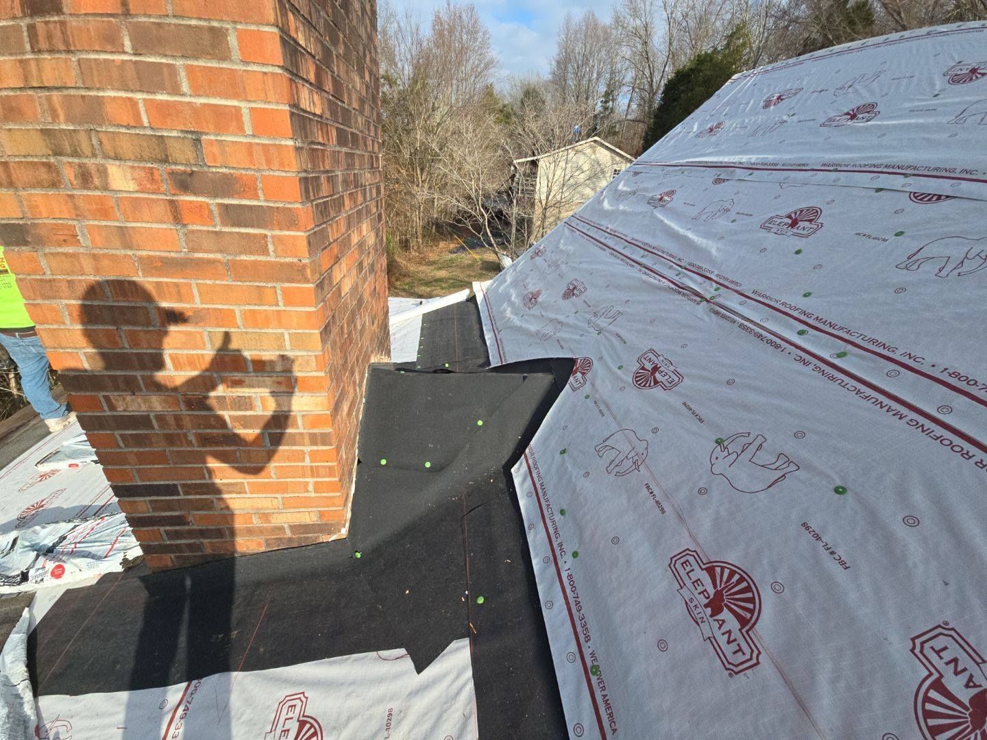 Roofer's shadow on roof with exposed underlayment next to a brick chimney; sunny day.