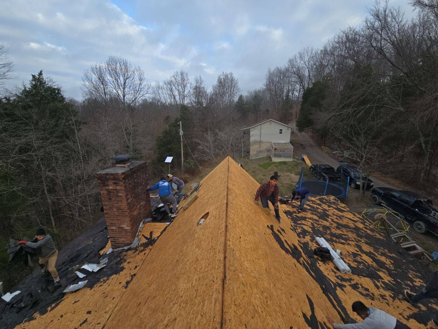 Roofers working on a house roof with exposed wood. Trees, sky, and a brick chimney are in the background.