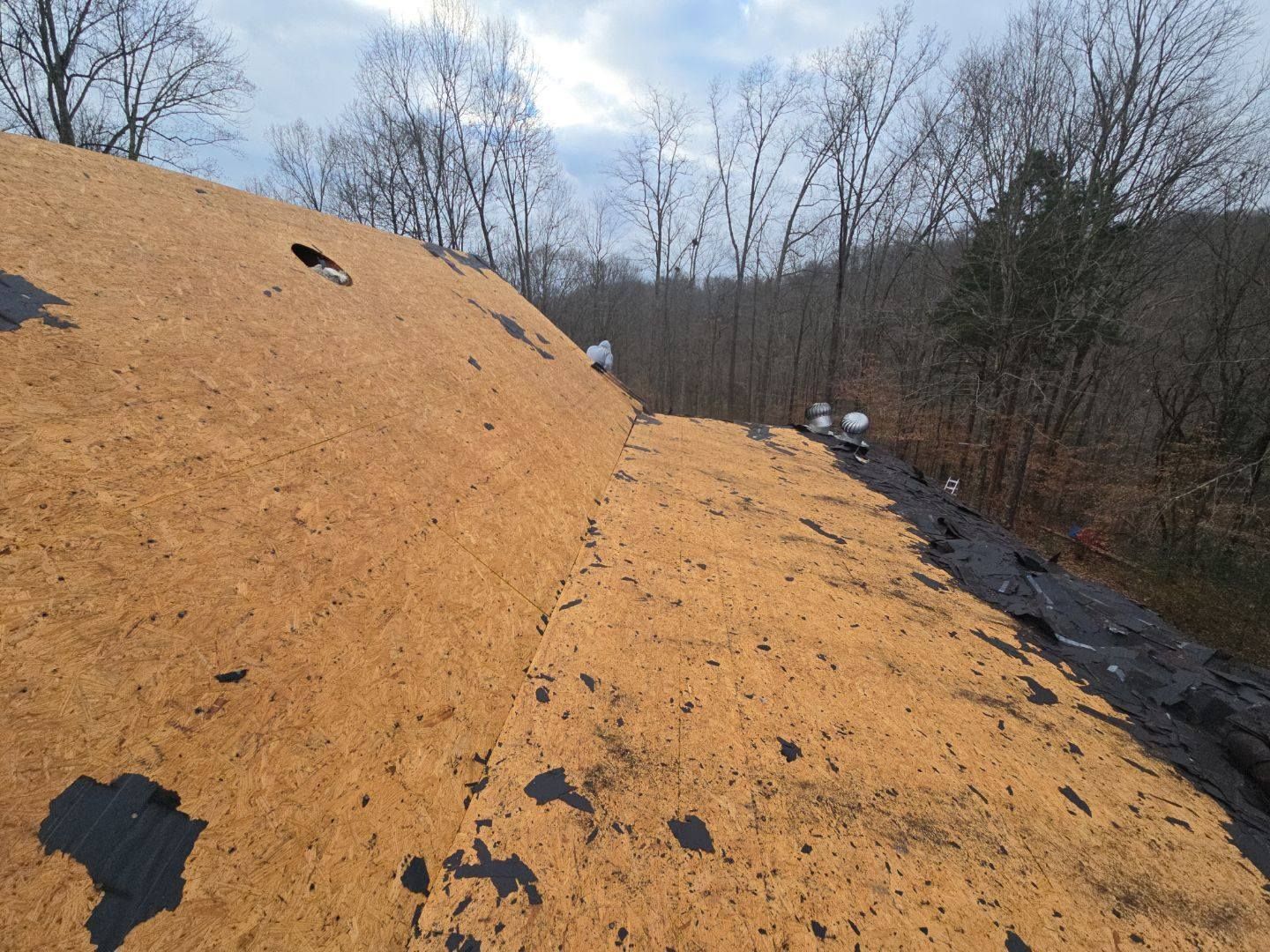 Rooftop with exposed brown sheathing and tear-out areas, in front of a tree-filled landscape.