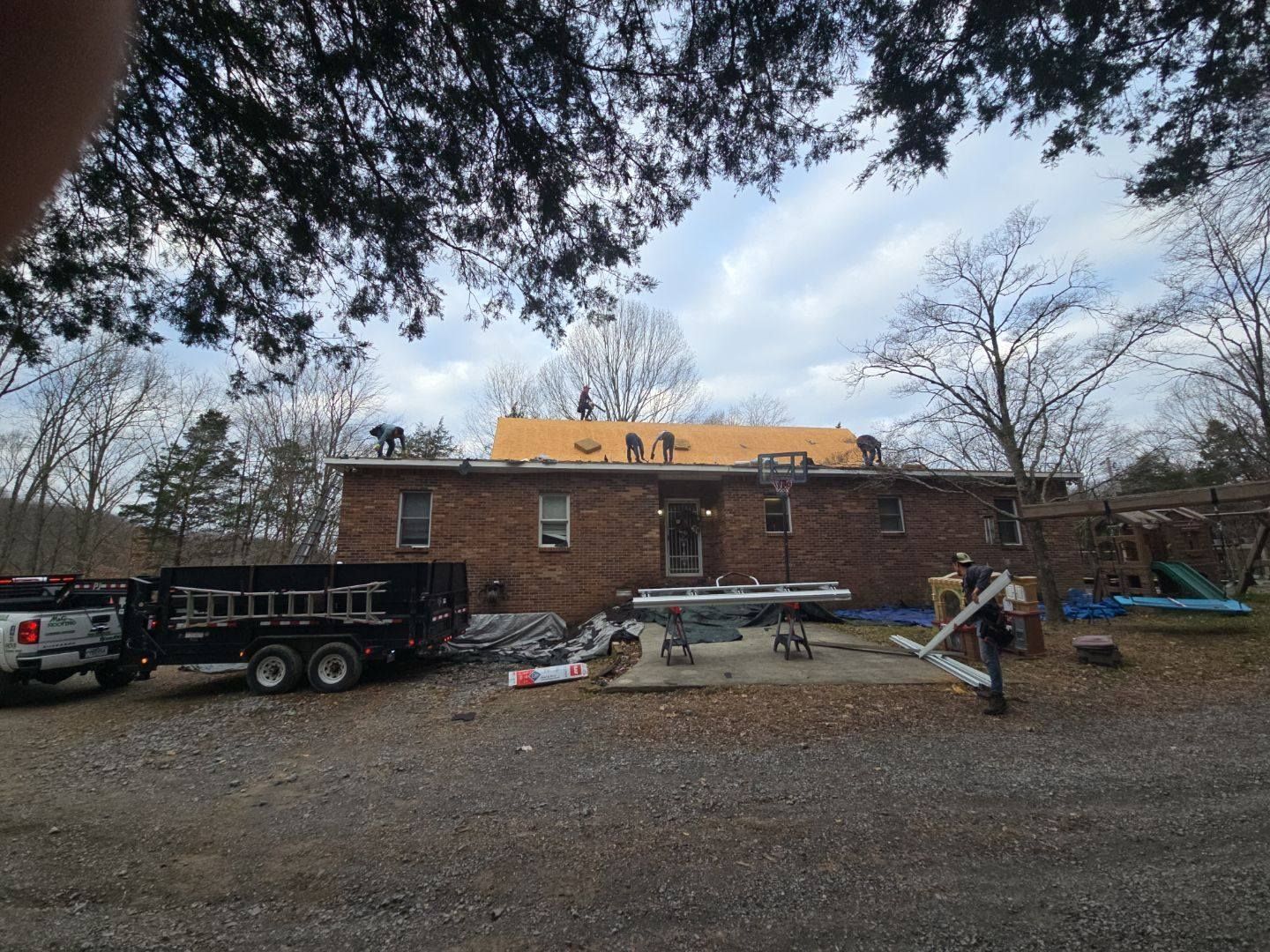 Workers on a brick house roof replacing shingles. Equipment and supplies are on the ground.
