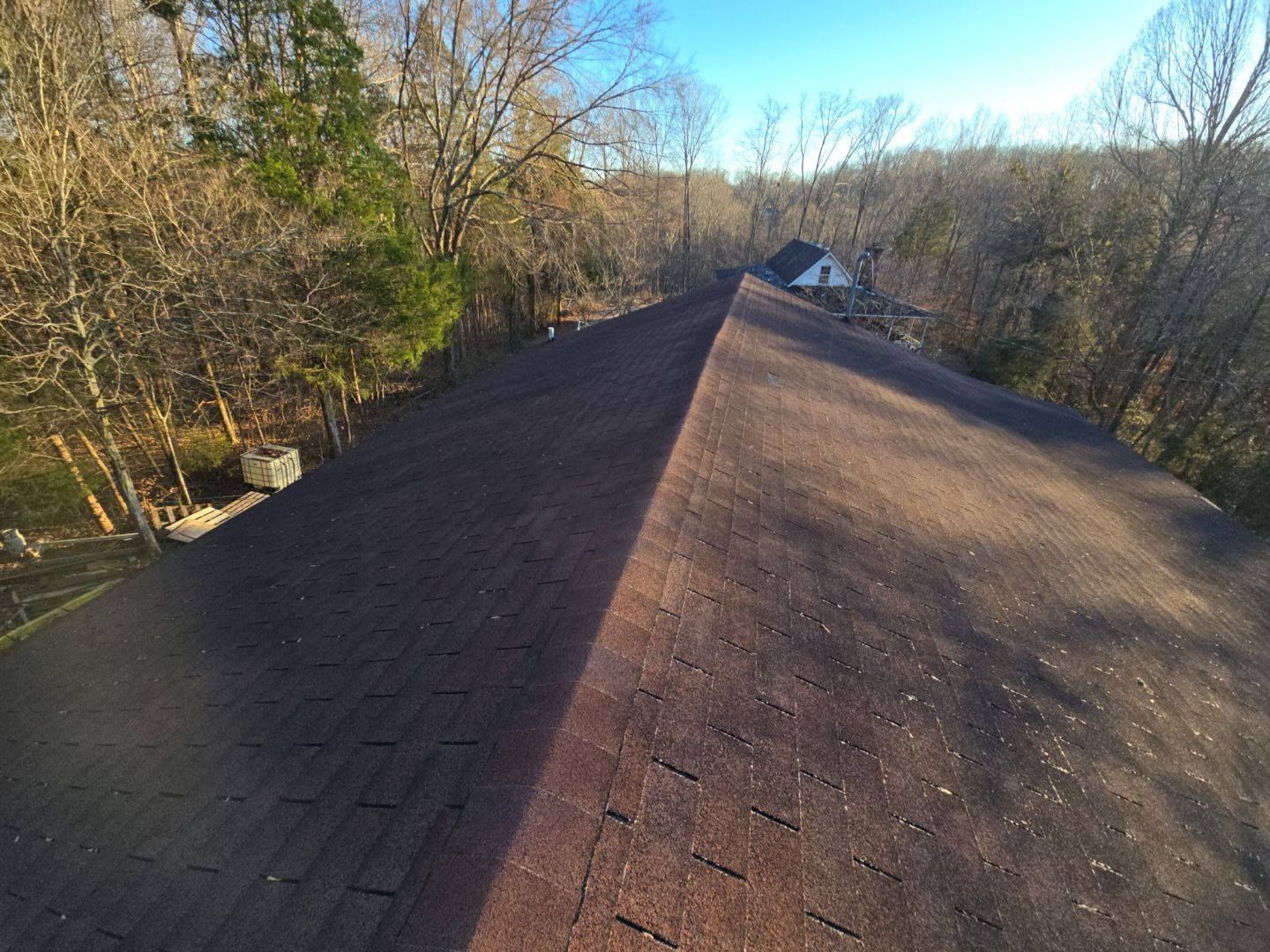 Brown asphalt shingle roof on a house, angled view, surrounded by trees under a clear blue sky.