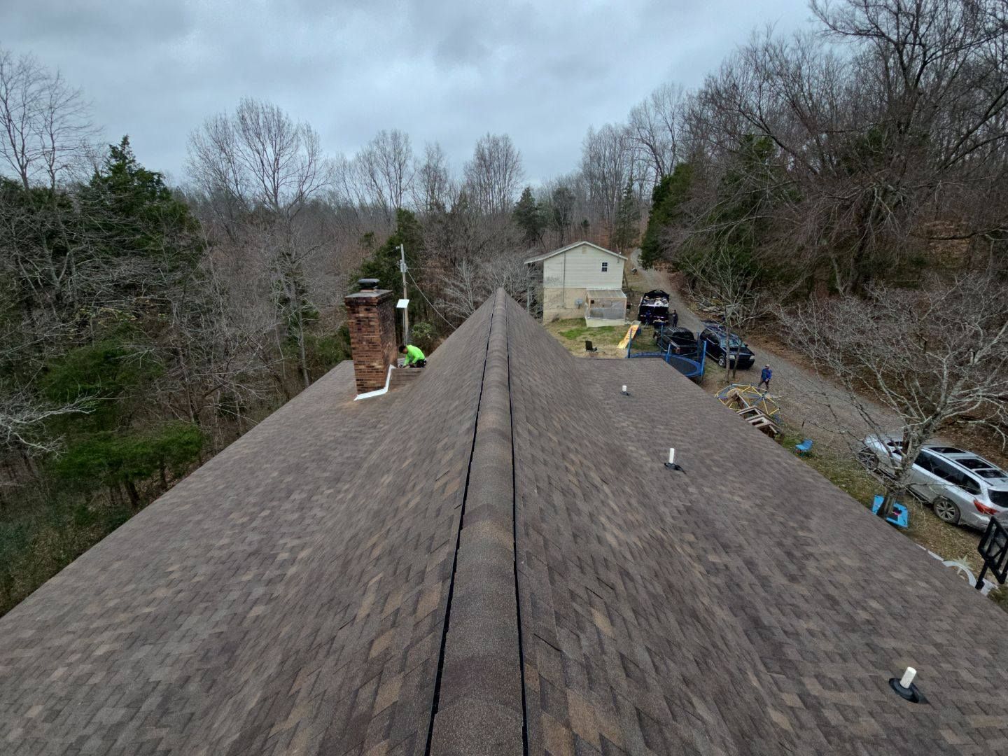 Brown shingled roof with a central ridge, chimney, and surrounding trees; vehicles parked in the background.