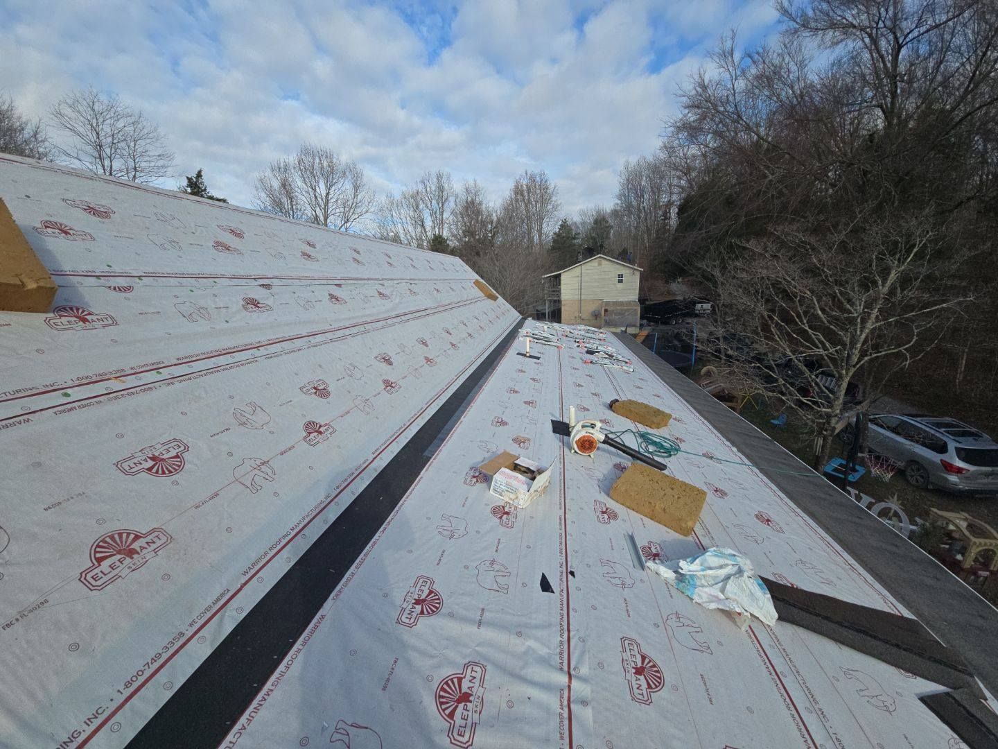 Rooftop with white underlayment, black strips, and materials scattered. Cloudy sky, trees, and a house in the background.