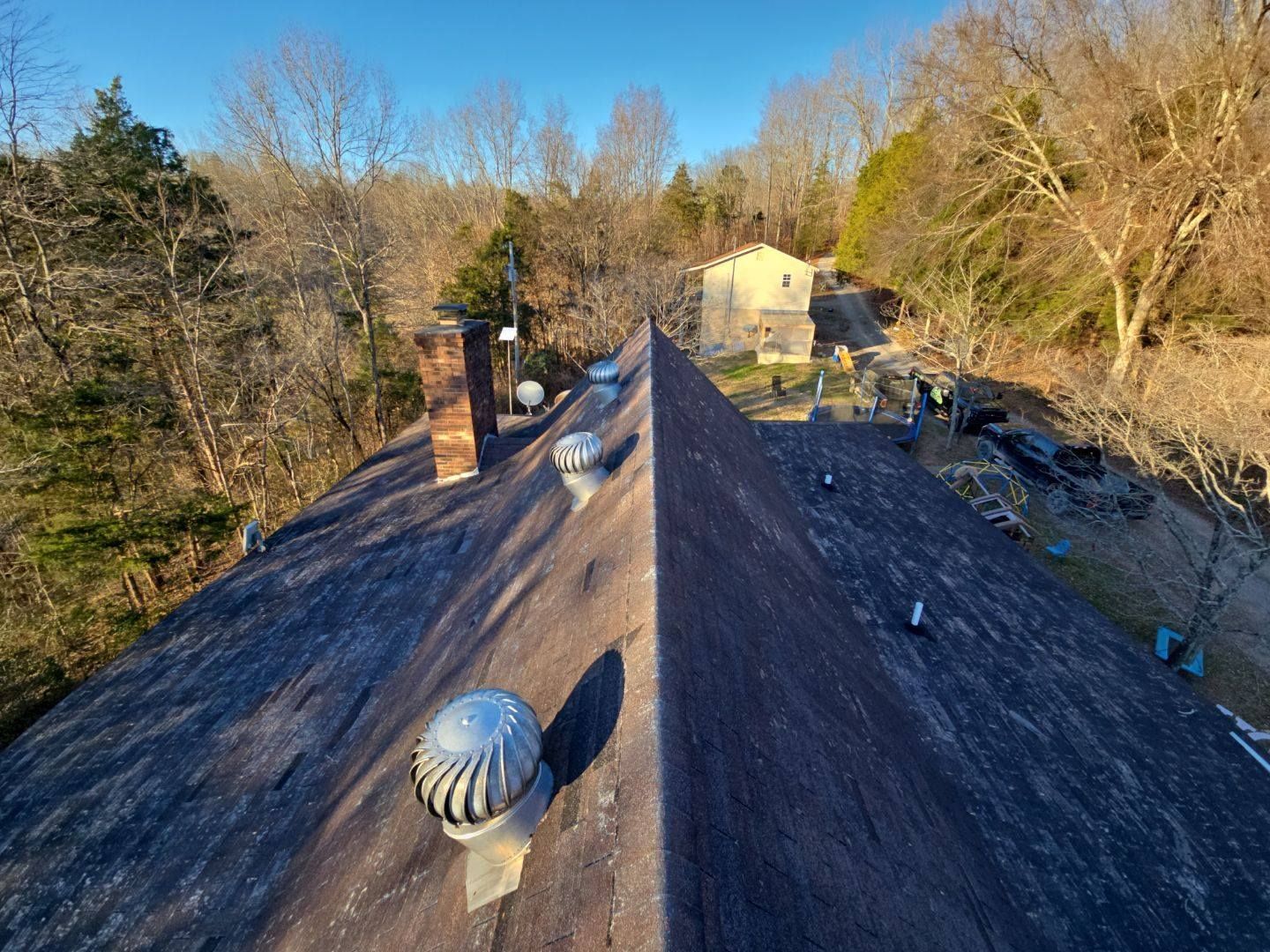 View from a roof, showing vents and a chimney, with a small building and trees in the distance on a sunny day.