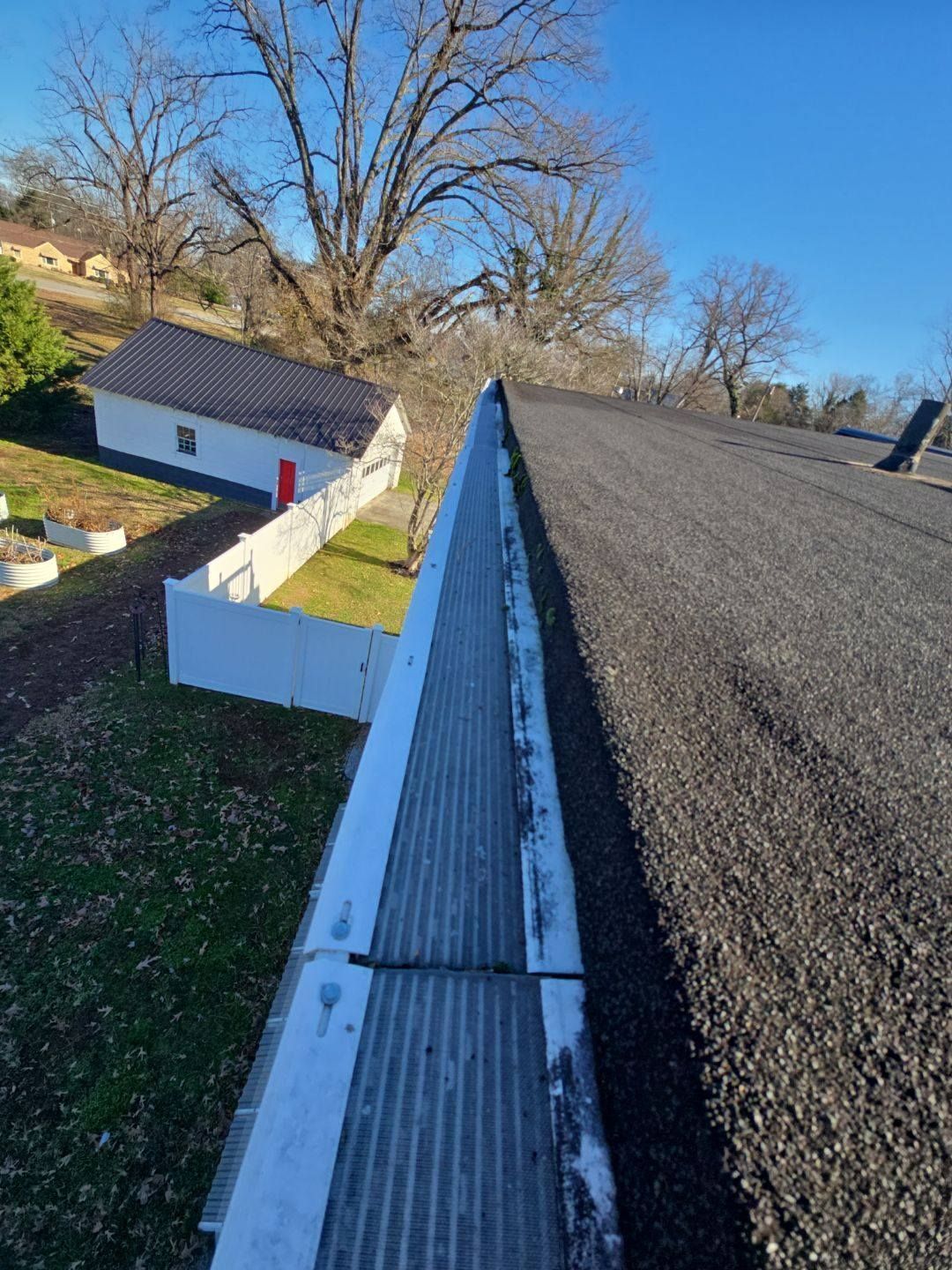 Rooftop view of gutter with debris and asphalt shingles, house and trees in background under a blue sky.