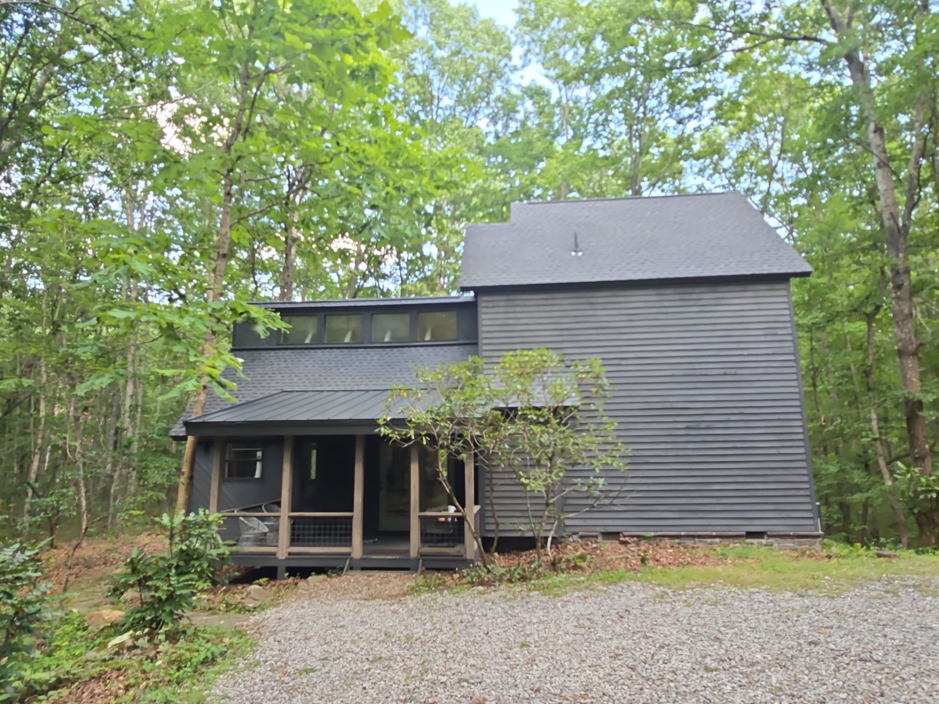 Dark gray cabin nestled in a forest. Porch with screening, gravel driveway. Trees surround the building.