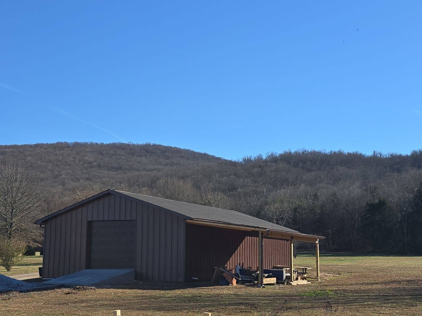 Brown barn with a covered porch in front of a tree-covered hillside on a sunny day.