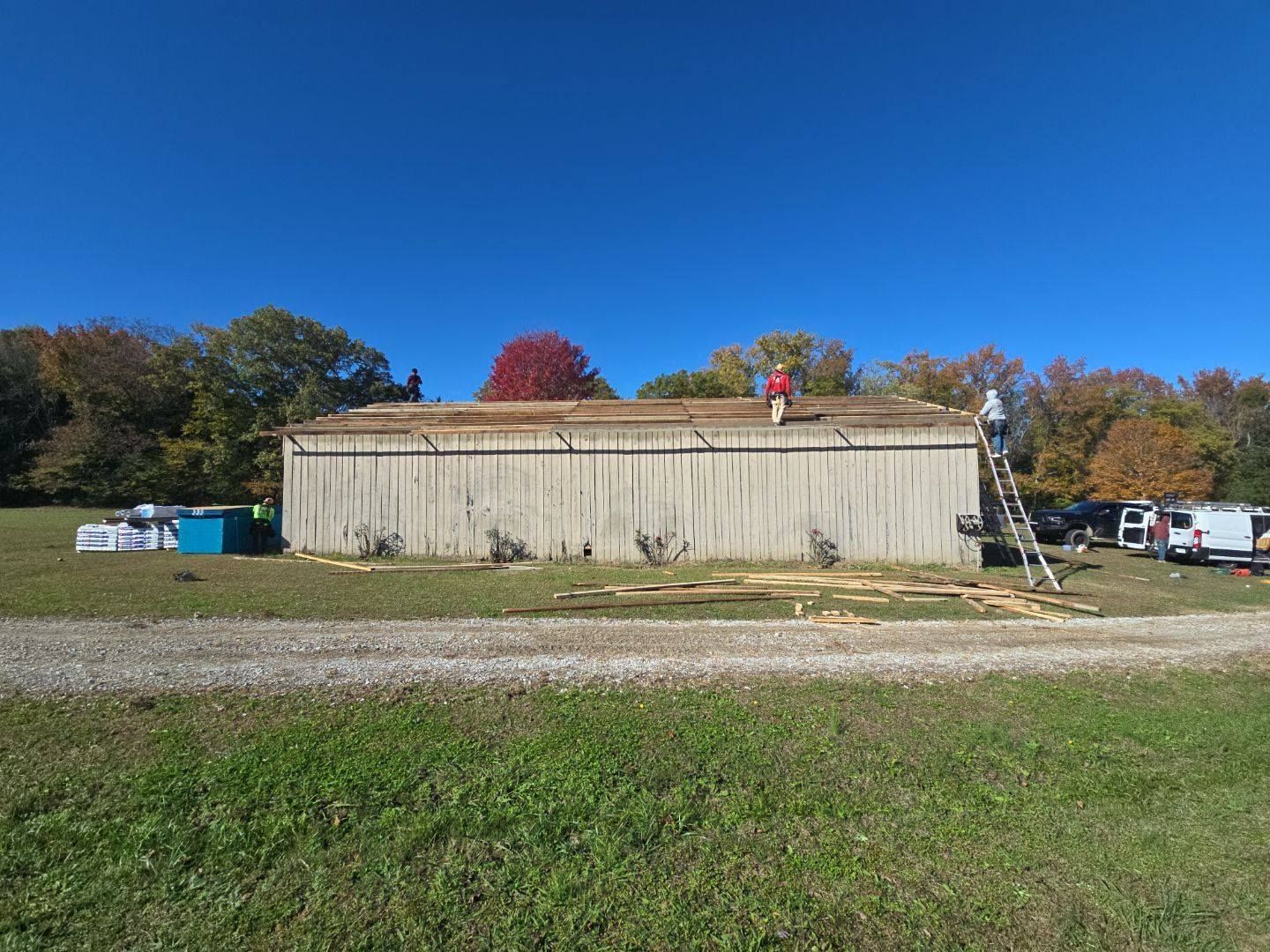 Workers on a roof, installing a new roof on a long, light-colored building in a grassy field with fall foliage.