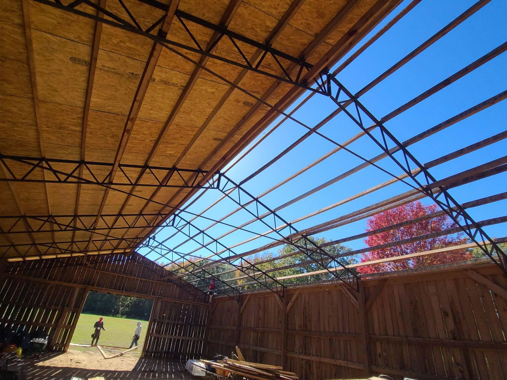 Interior view of a barn under construction with exposed wooden framework and blue sky peeking through the roof.