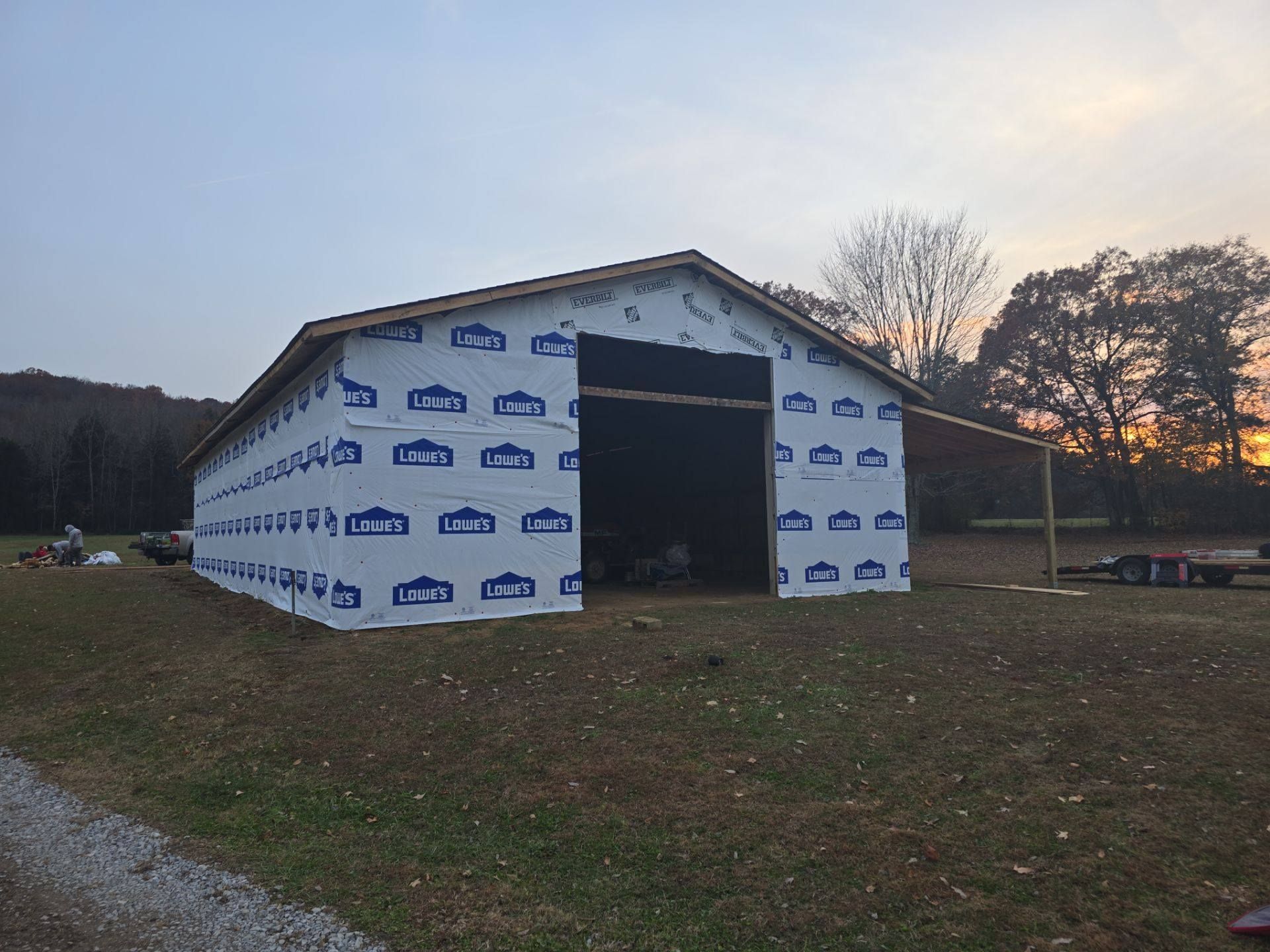 Barn under construction with blue and white wrap, open doorway, and a sunset in the background.