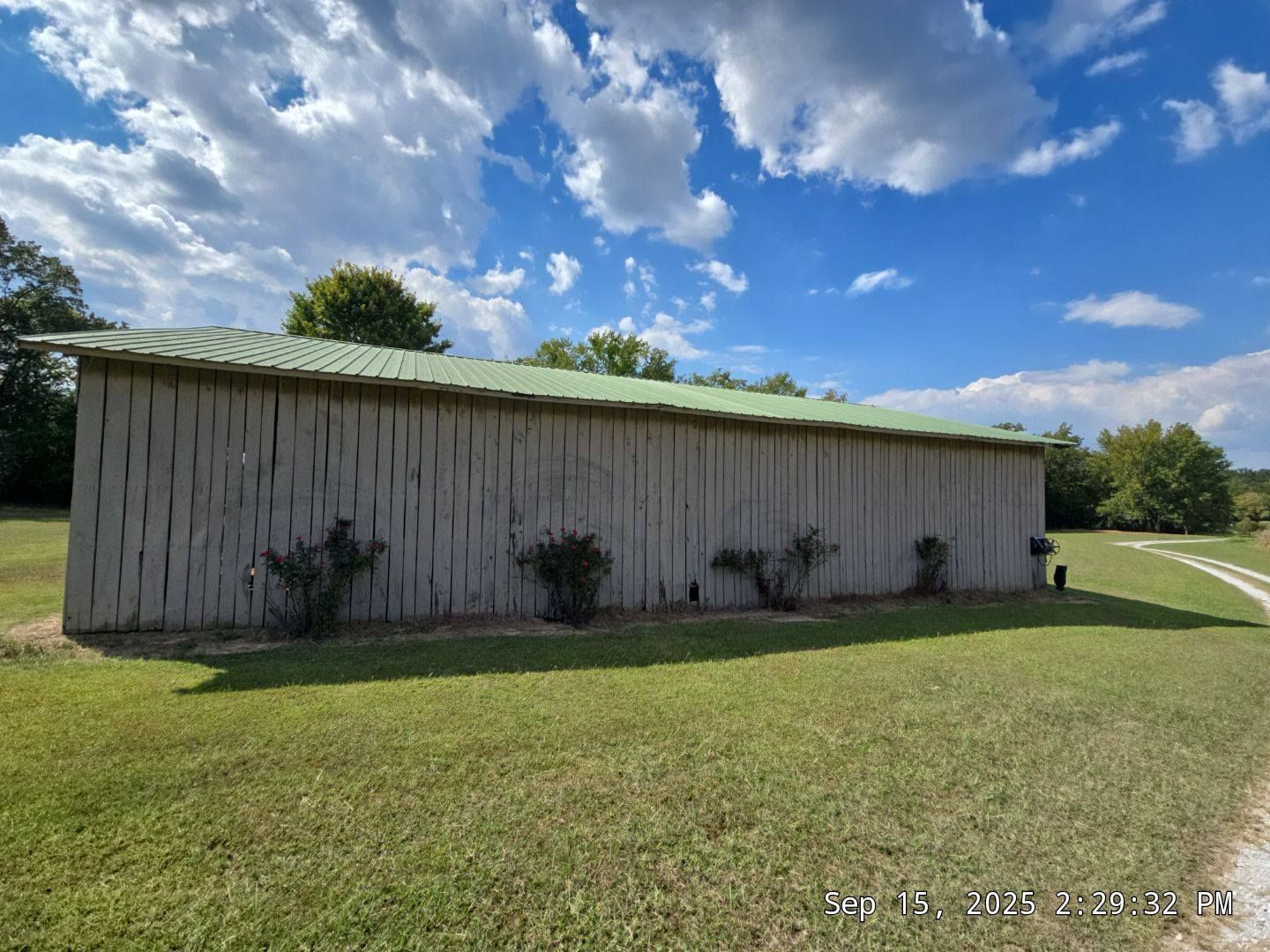 A long, weathered barn with a green roof sits in a grassy field under a cloudy blue sky.