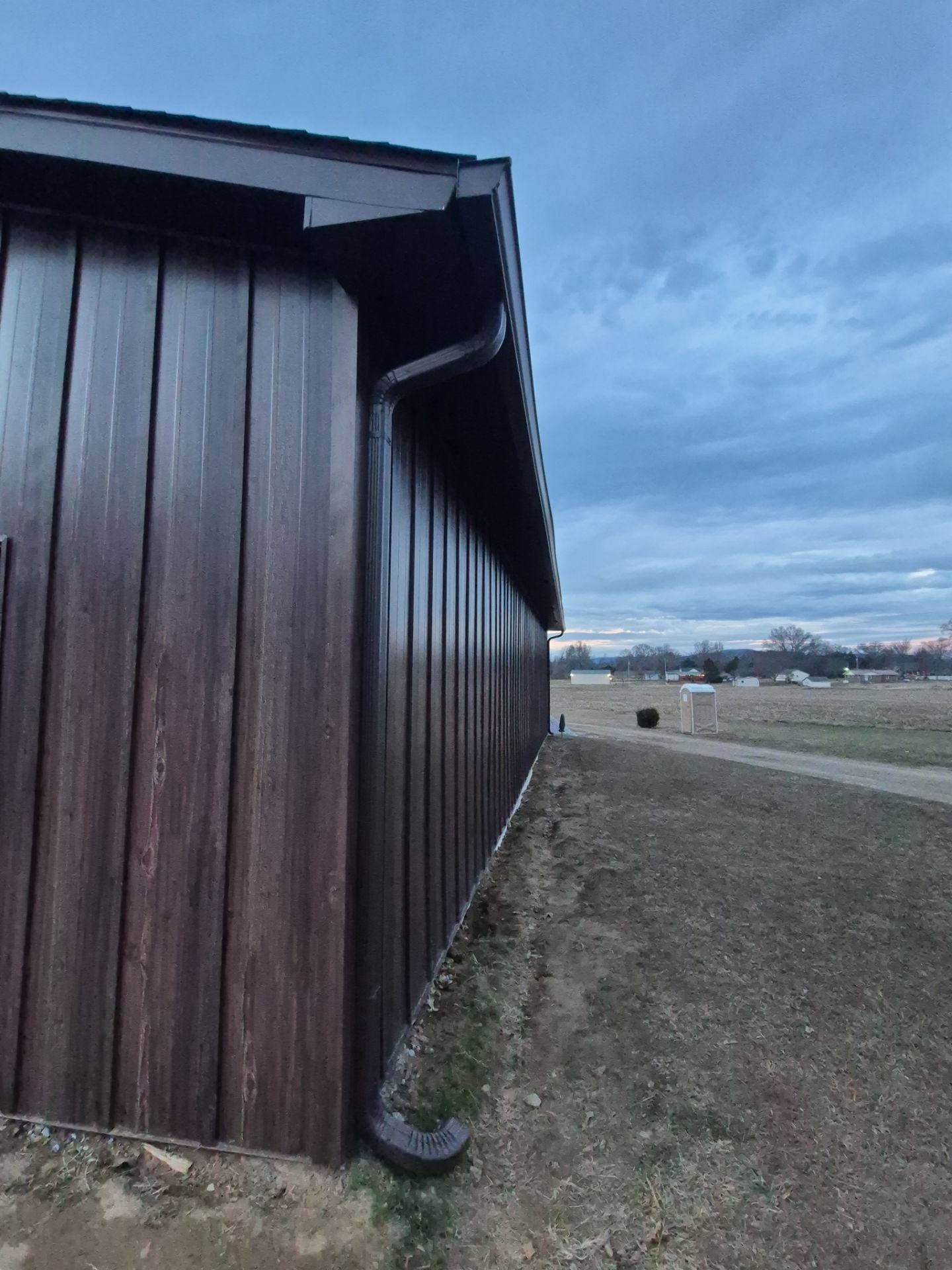 Brown metal-sided building with dark trim, gutter, and a cloudy sky background.