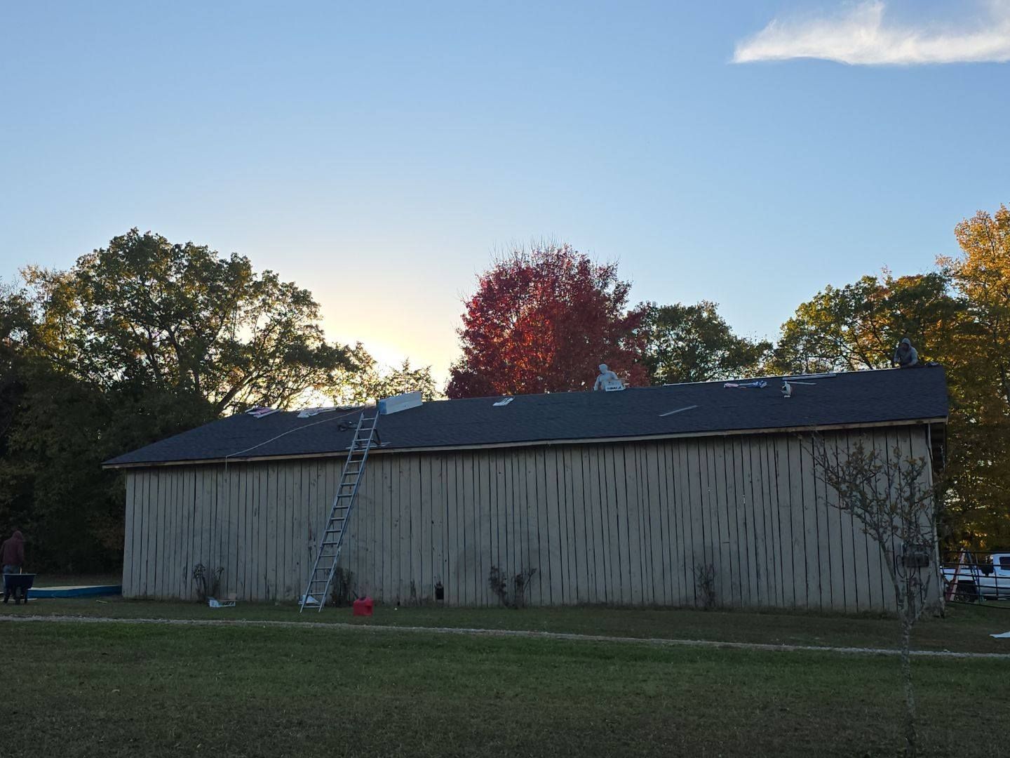 A long, light-brown building with a dark roof being worked on, ladder against the side, trees in background.