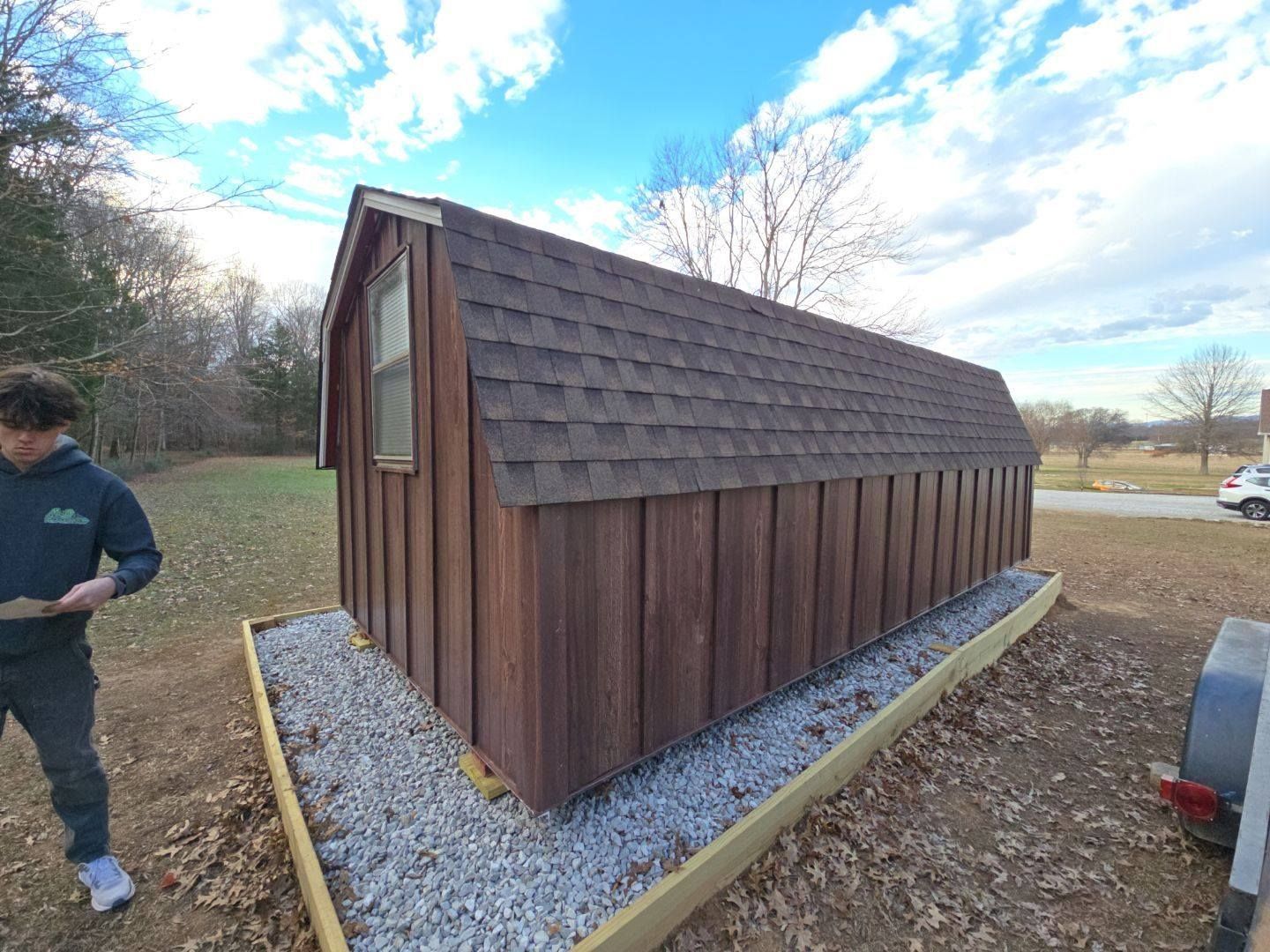 Brown shed with a gravel base, boy standing nearby, blue sky.