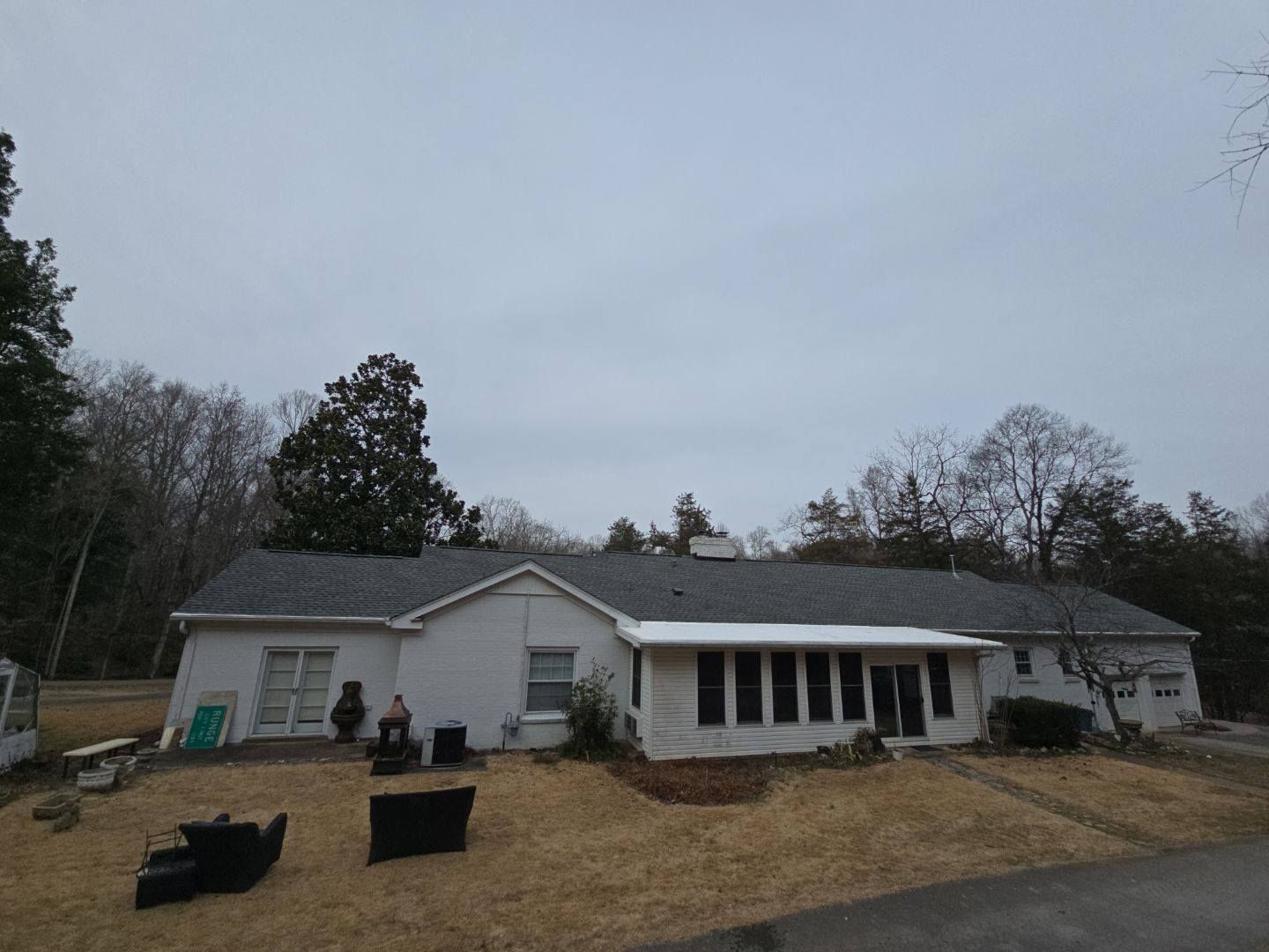 Single-story white house with a dark roof on a cloudy day, surrounded by trees and a brown yard.