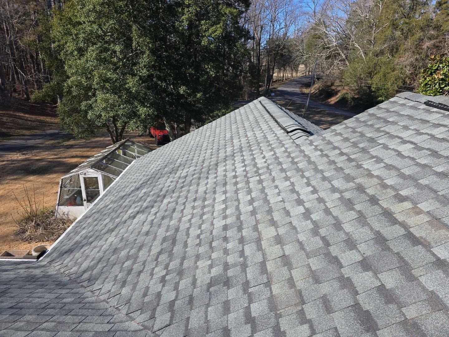 Gray asphalt shingle roof on a house, angled view, with a treeline and greenhouse visible in the background.