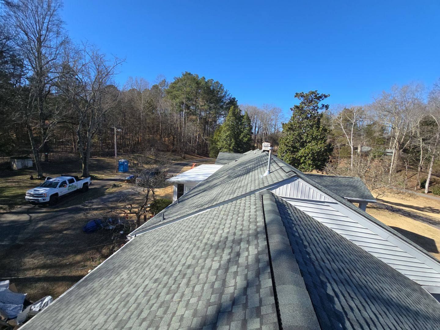 Rooftop view of gray shingle roof, chimney, trees, clear blue sky, and a white truck in the yard.