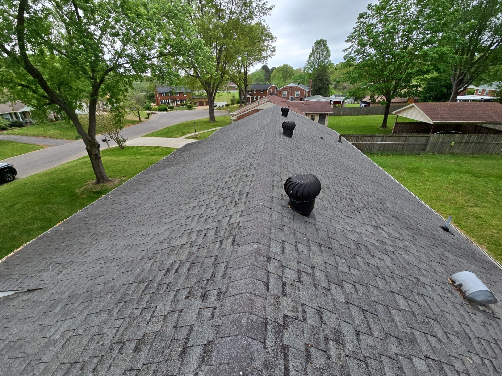 View of a residential shingle roof with vents, overlooking a neighborhood with trees and houses on a cloudy day.