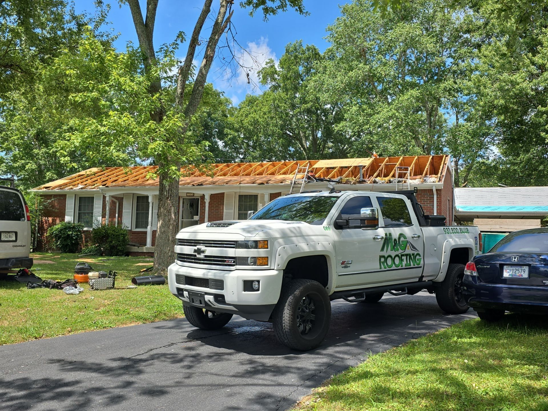 White pickup truck with company logo parked in front of house undergoing roof work. Trees and blue sky are in the background.