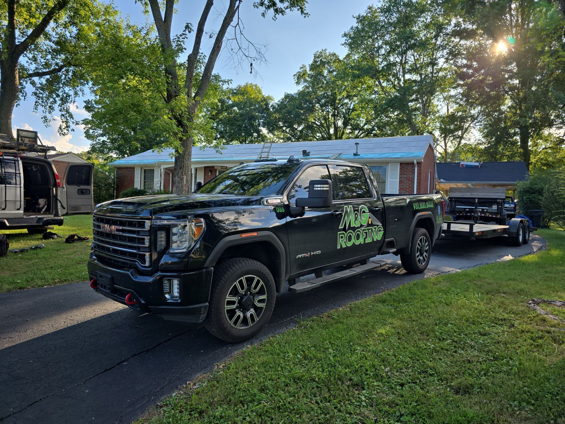 Black GMC truck with trailer parked in a driveway, house in the background. Green company logo on the truck.
