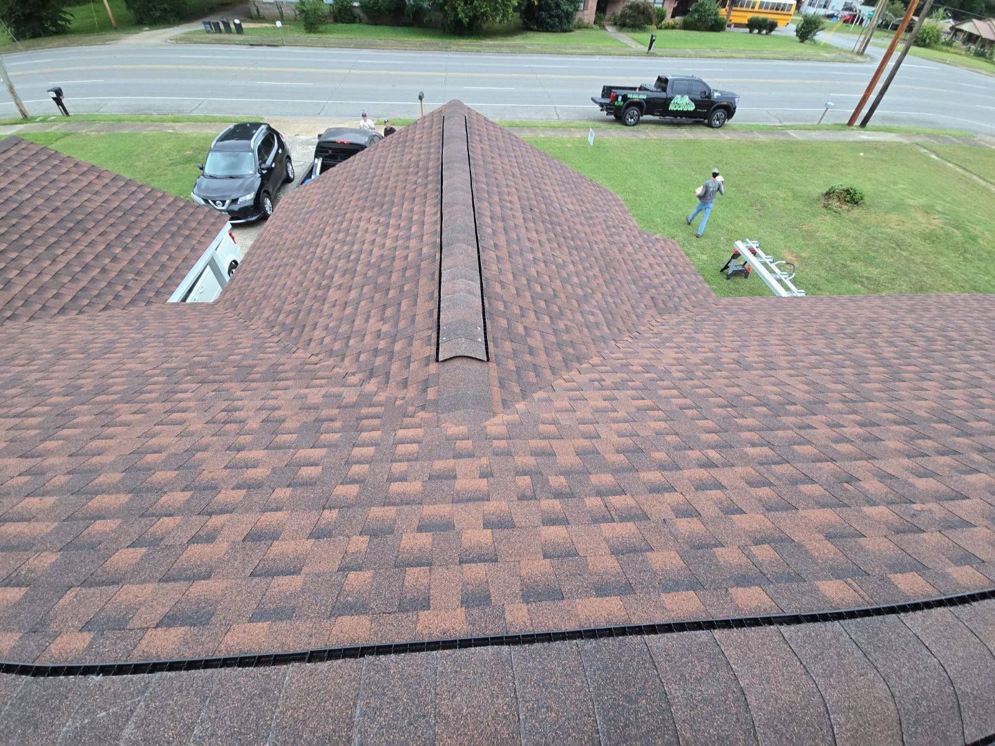 Brown shingled roof with a dark ridge, overlooking a street and yard where a person works.