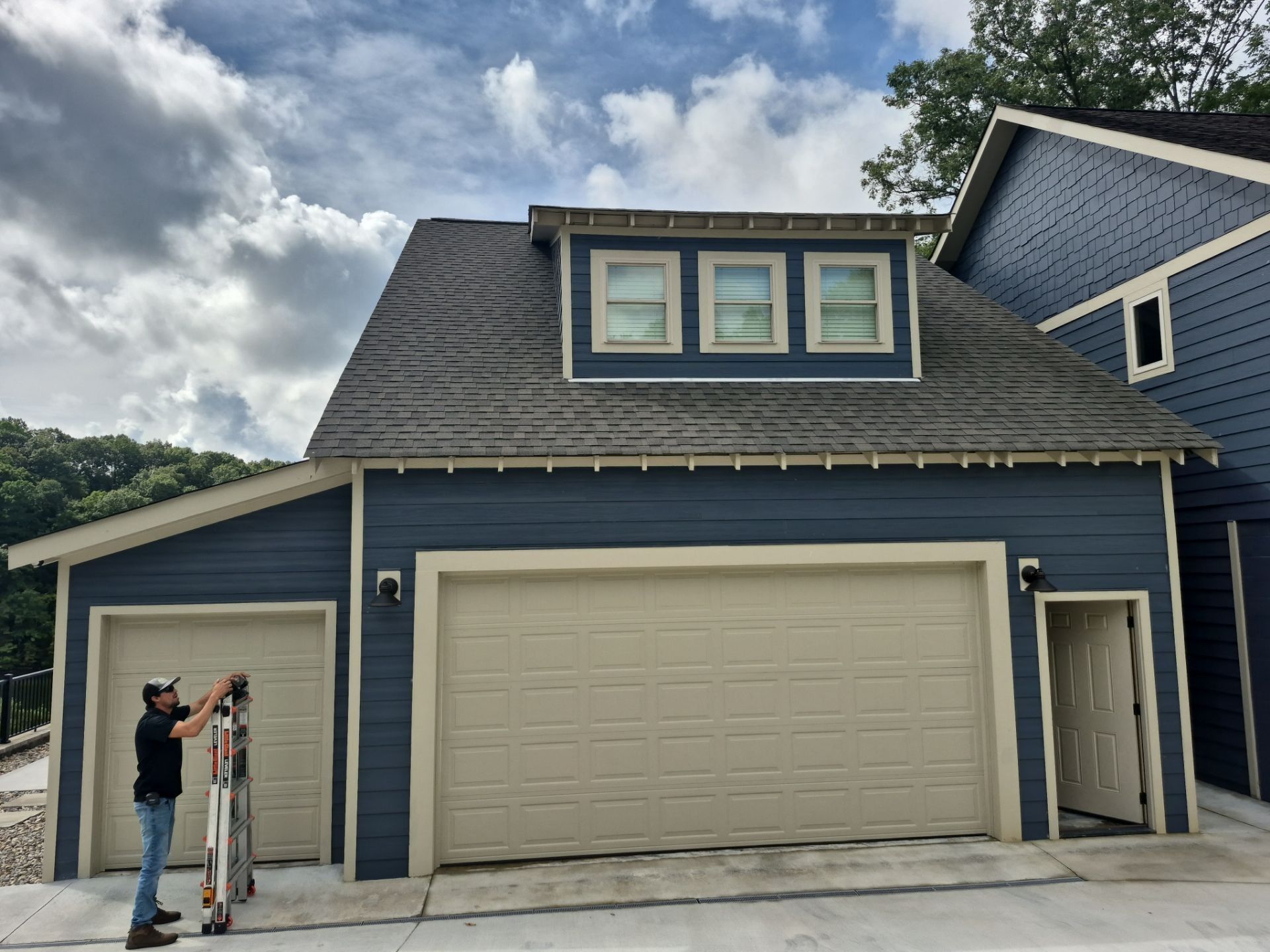 Blue garage with tan doors, a dormer, and a person using a ladder.