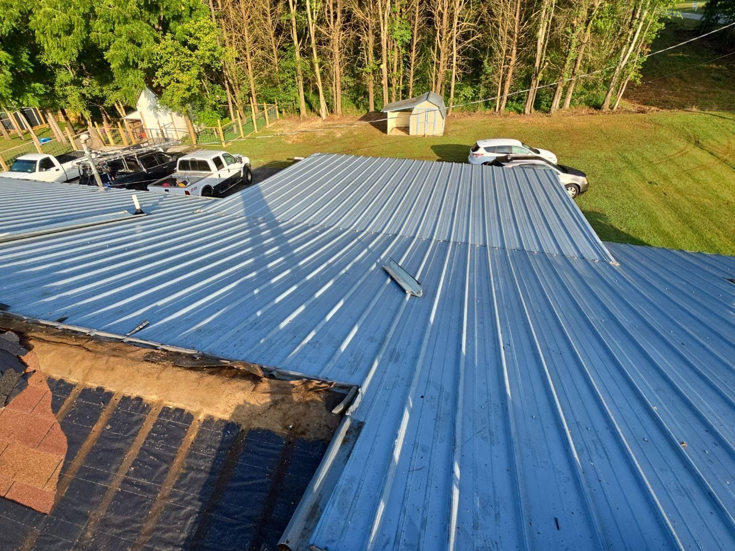 A blue metal roof on a building with multiple vehicles parked nearby, surrounded by trees and grass.