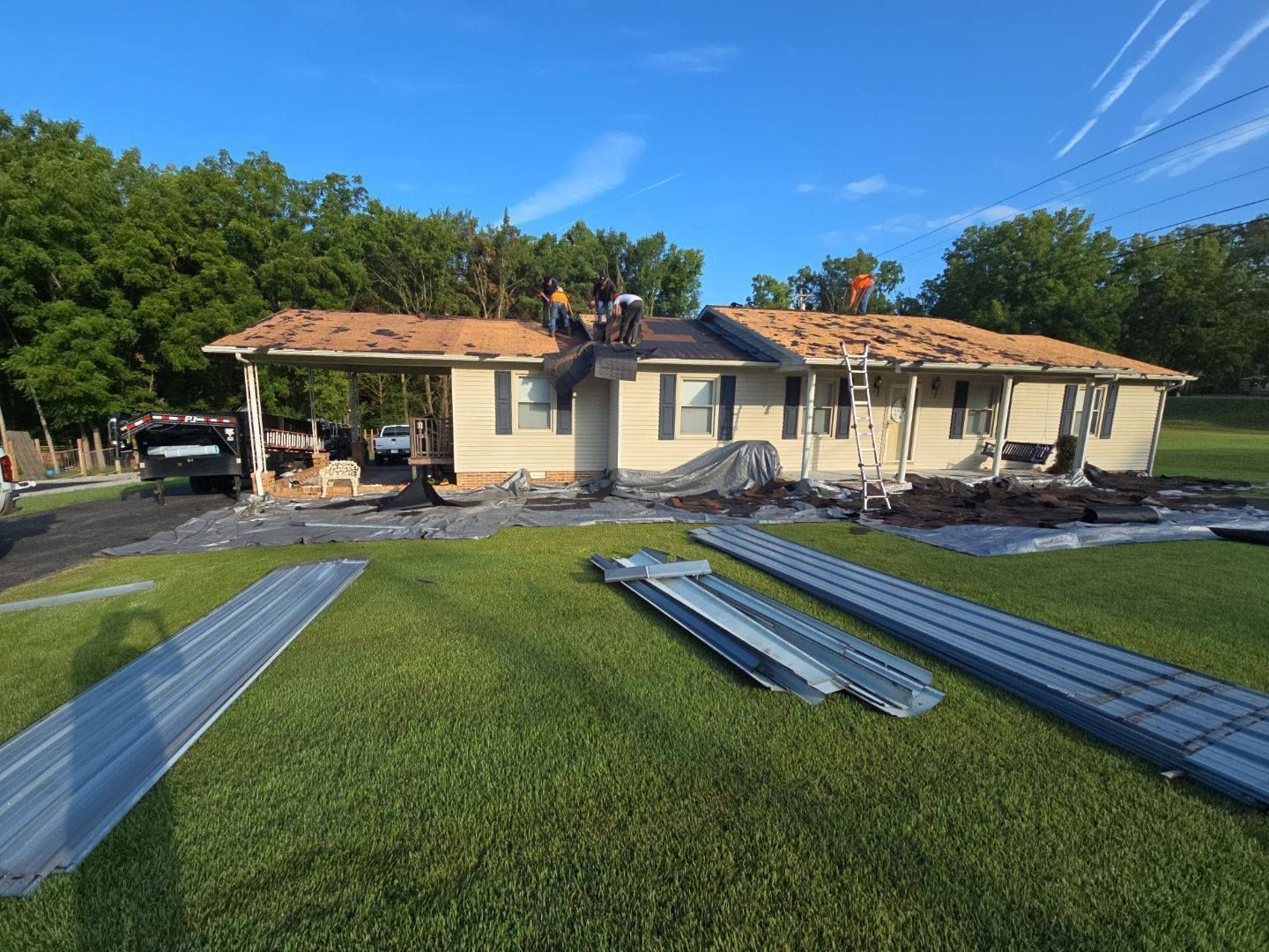 Roofers installing a metal roof on a single-story house. Materials and workers visible; green lawn.