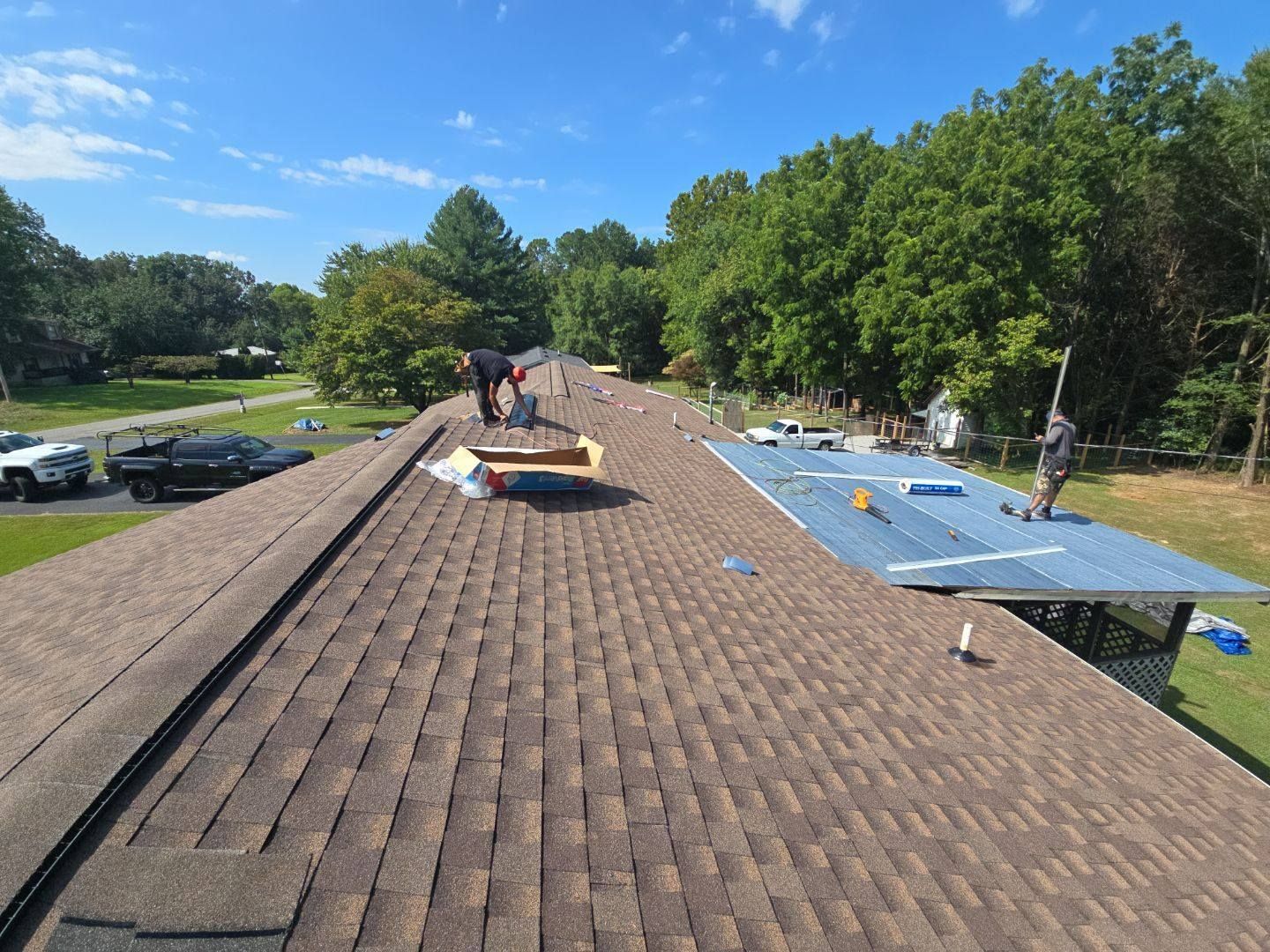 Roofers working on a brown shingle roof on a sunny day. Trees in the background.