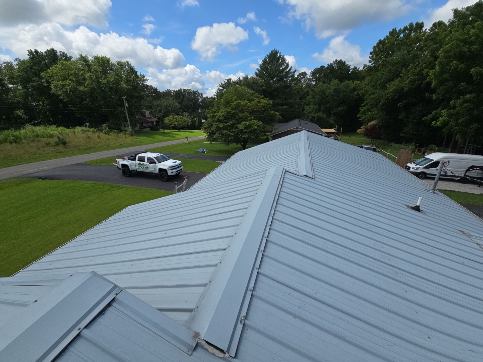Metal roof of a building with a white truck on a driveway below, surrounded by trees.