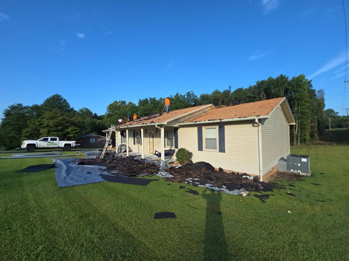 House with roofing work in progress; workers on the roof, debris on the ground, and a truck parked on the grass.
