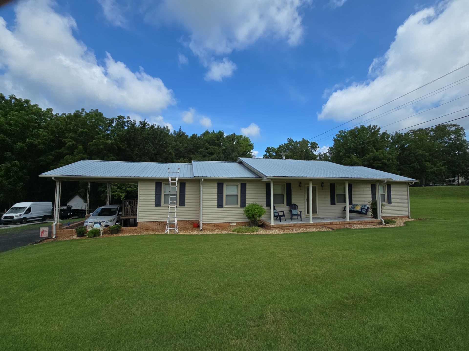 Beige one-story house with blue metal roof, green lawn, trees, and cloudy sky. Carport on the left.
