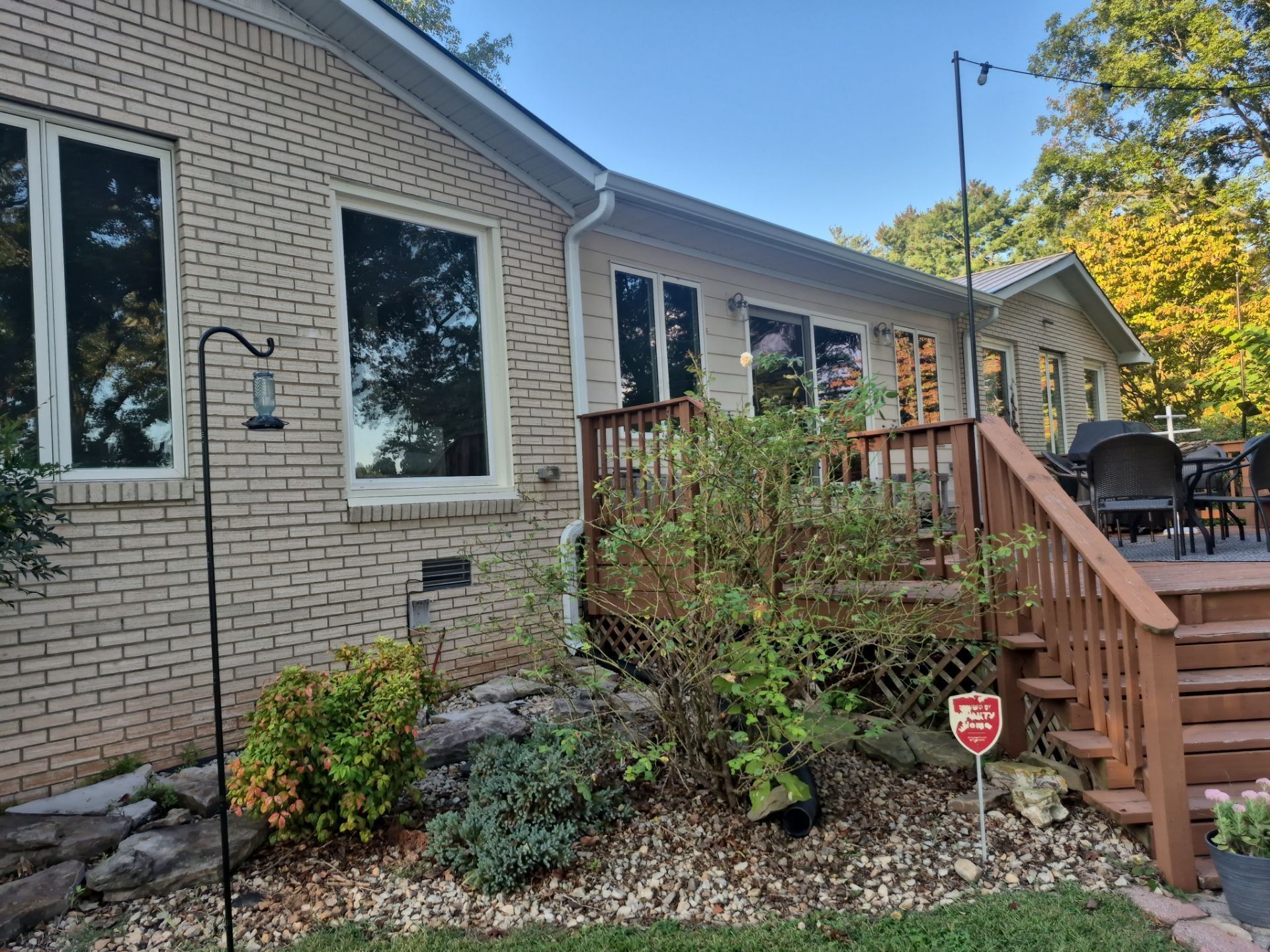 Beige brick house with deck, landscaping, windows, and security system sign.