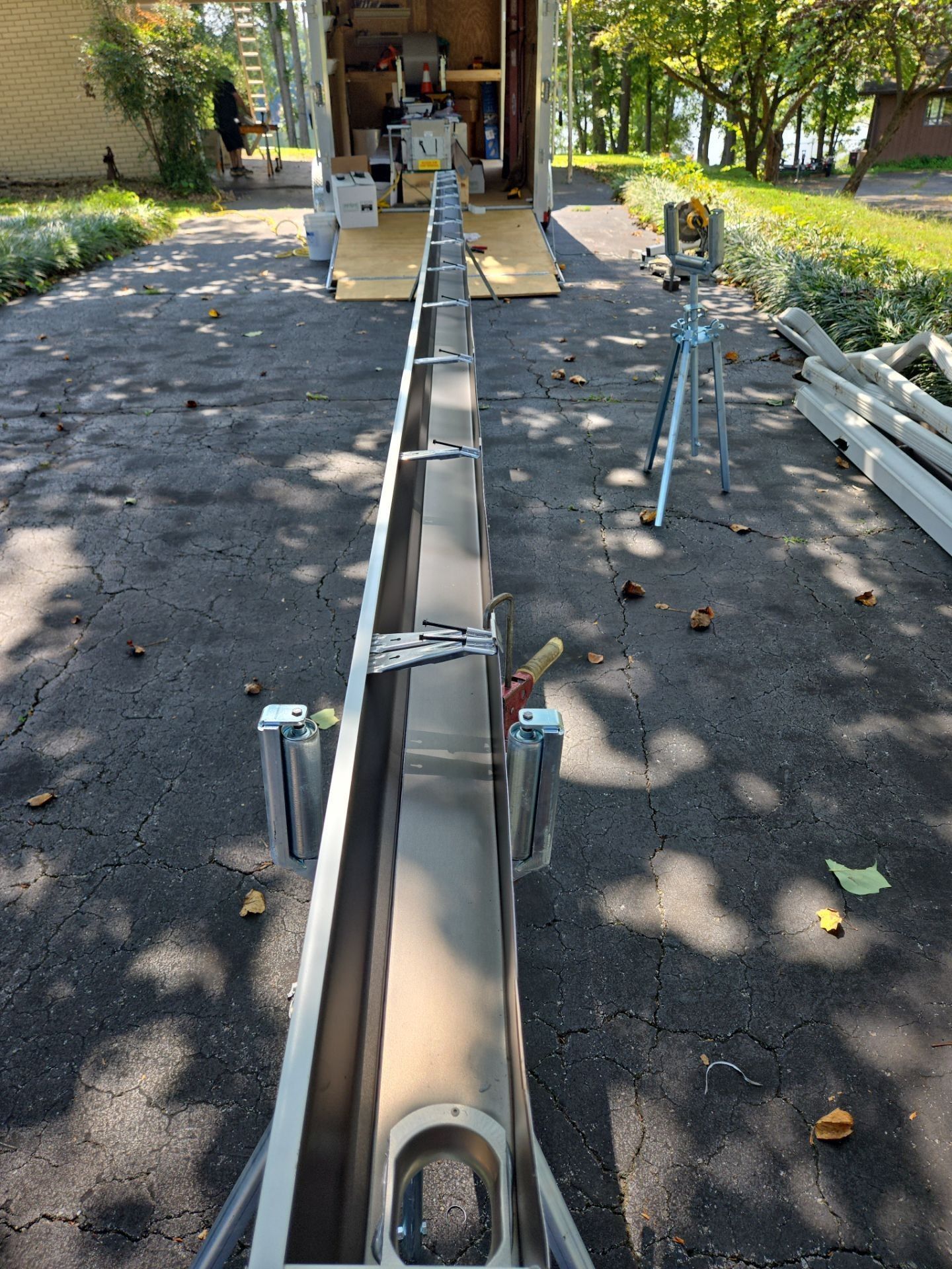 Long metal gutter on supports, tools, and a trailer on asphalt driveway.