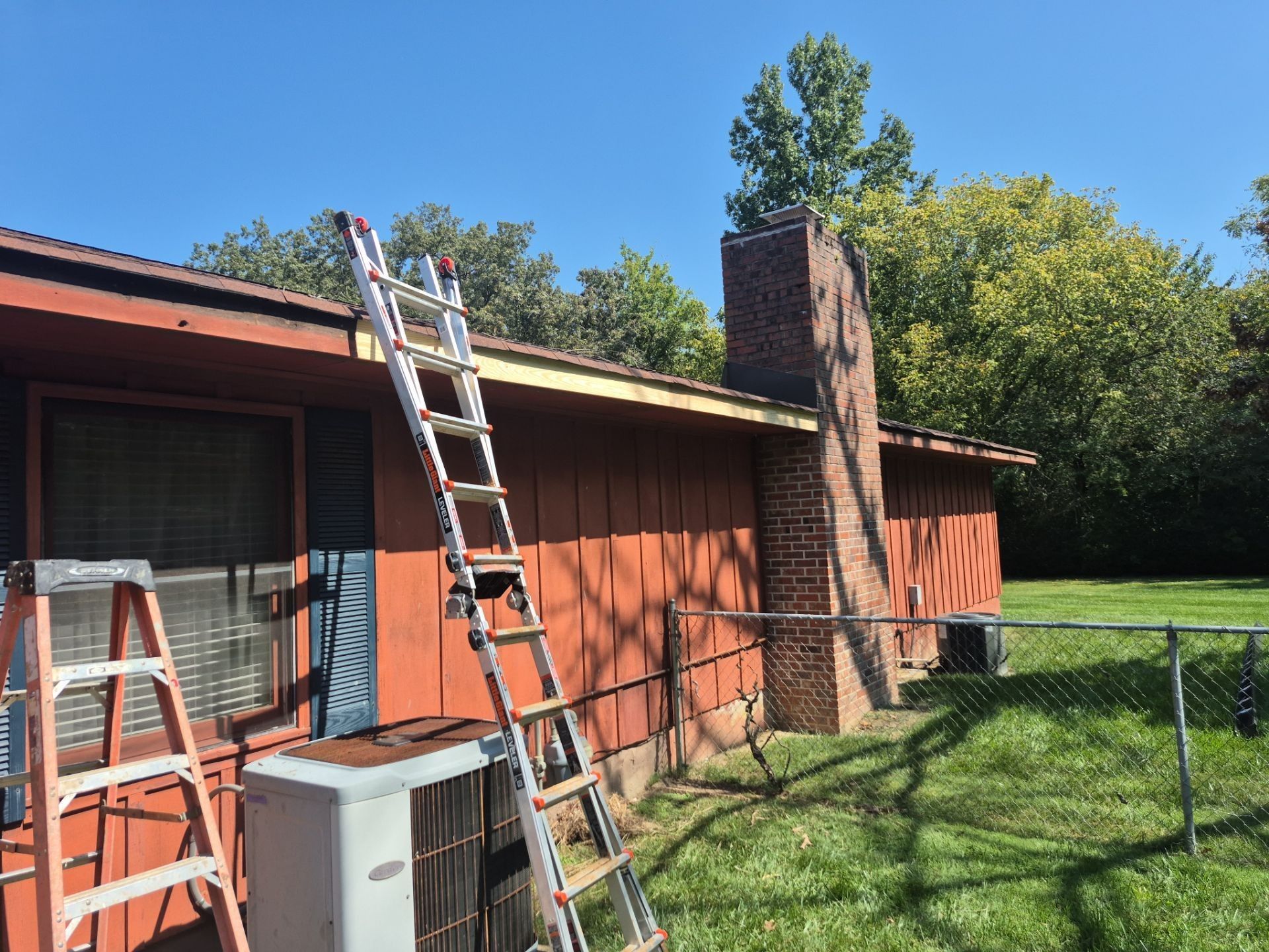 Ladder leaning against a red-sided house with a brick chimney on a sunny day.