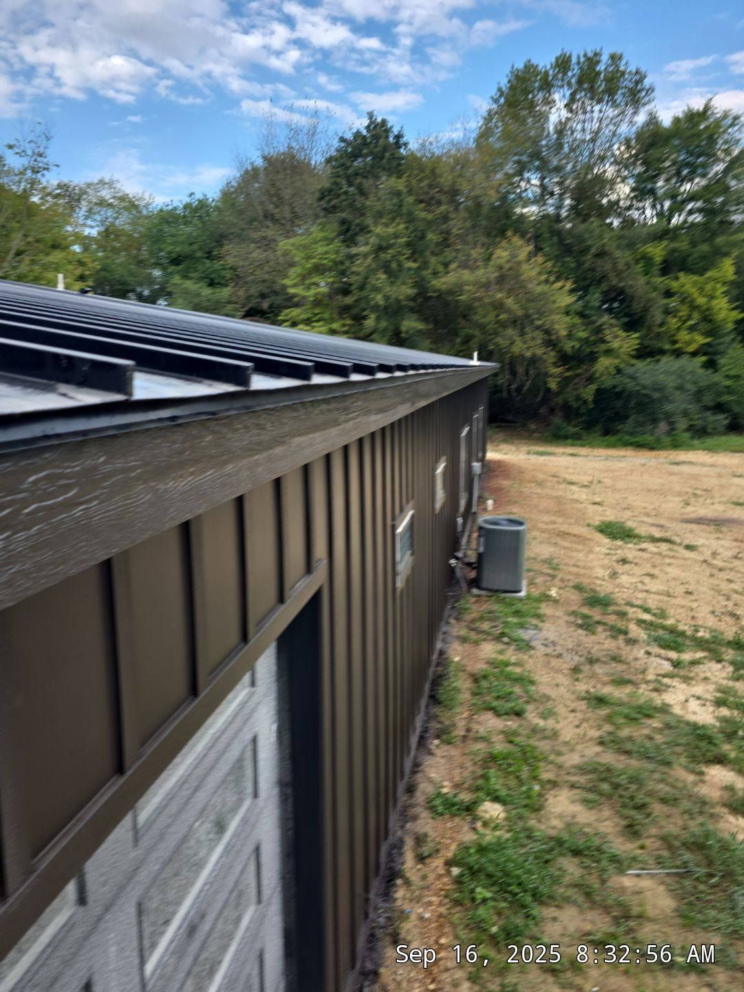 Brown metal-sided building with black roof and white door, set against a backdrop of trees and a blue sky.
