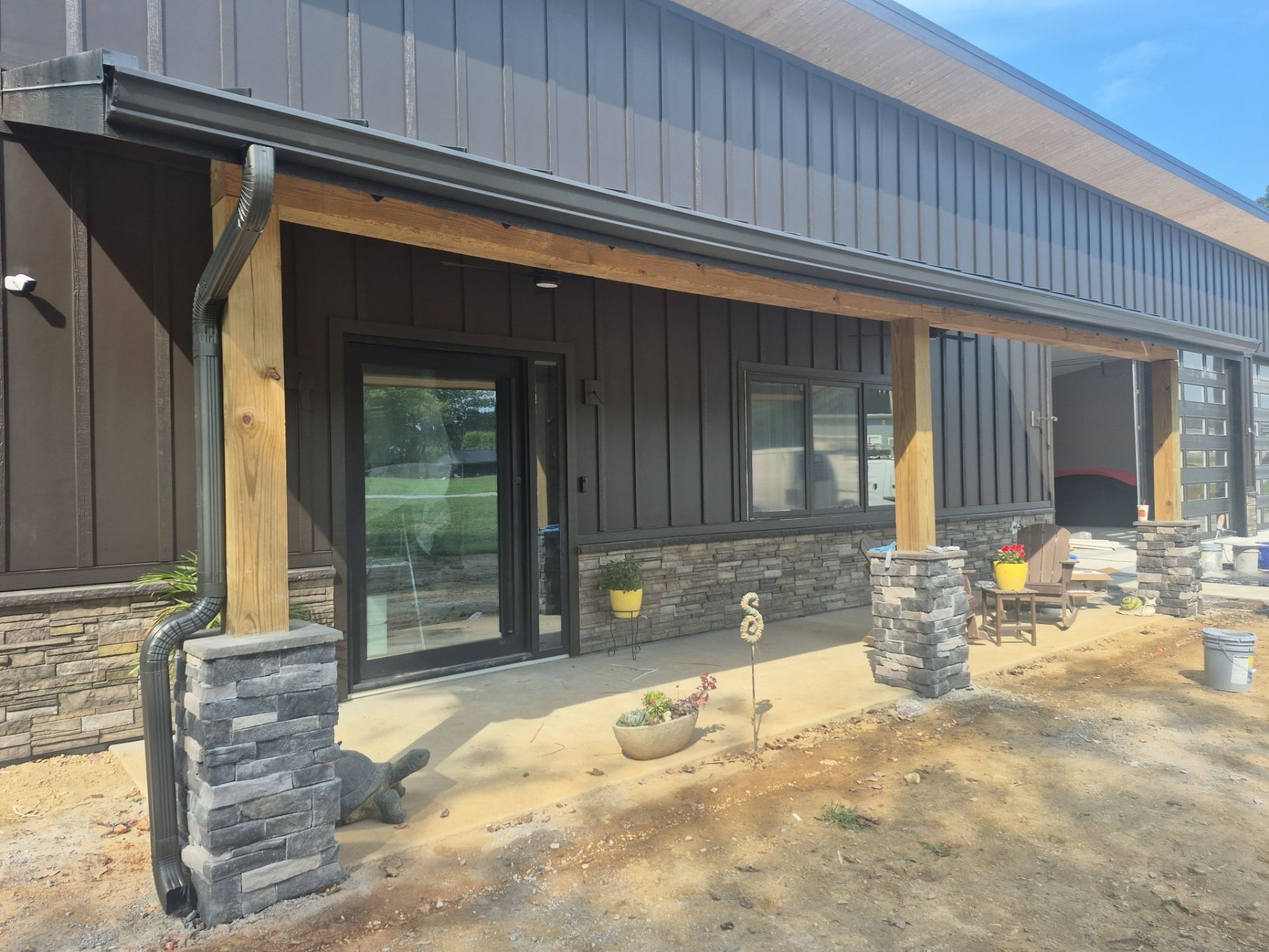 Covered porch with dark siding and stone-wrapped columns. Glass door and window. Sunny day.
