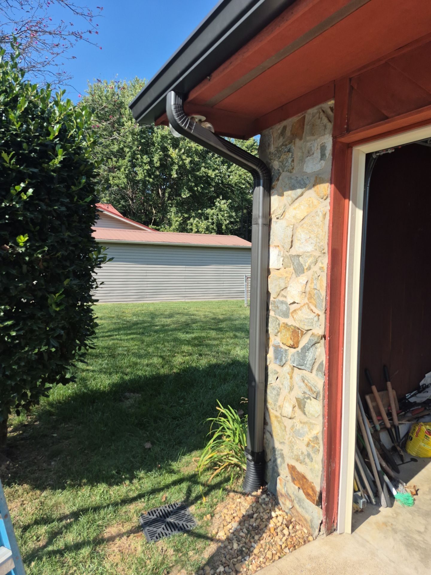 Black rain gutter on a stone house corner, with green lawn and shrubs, blue sky.