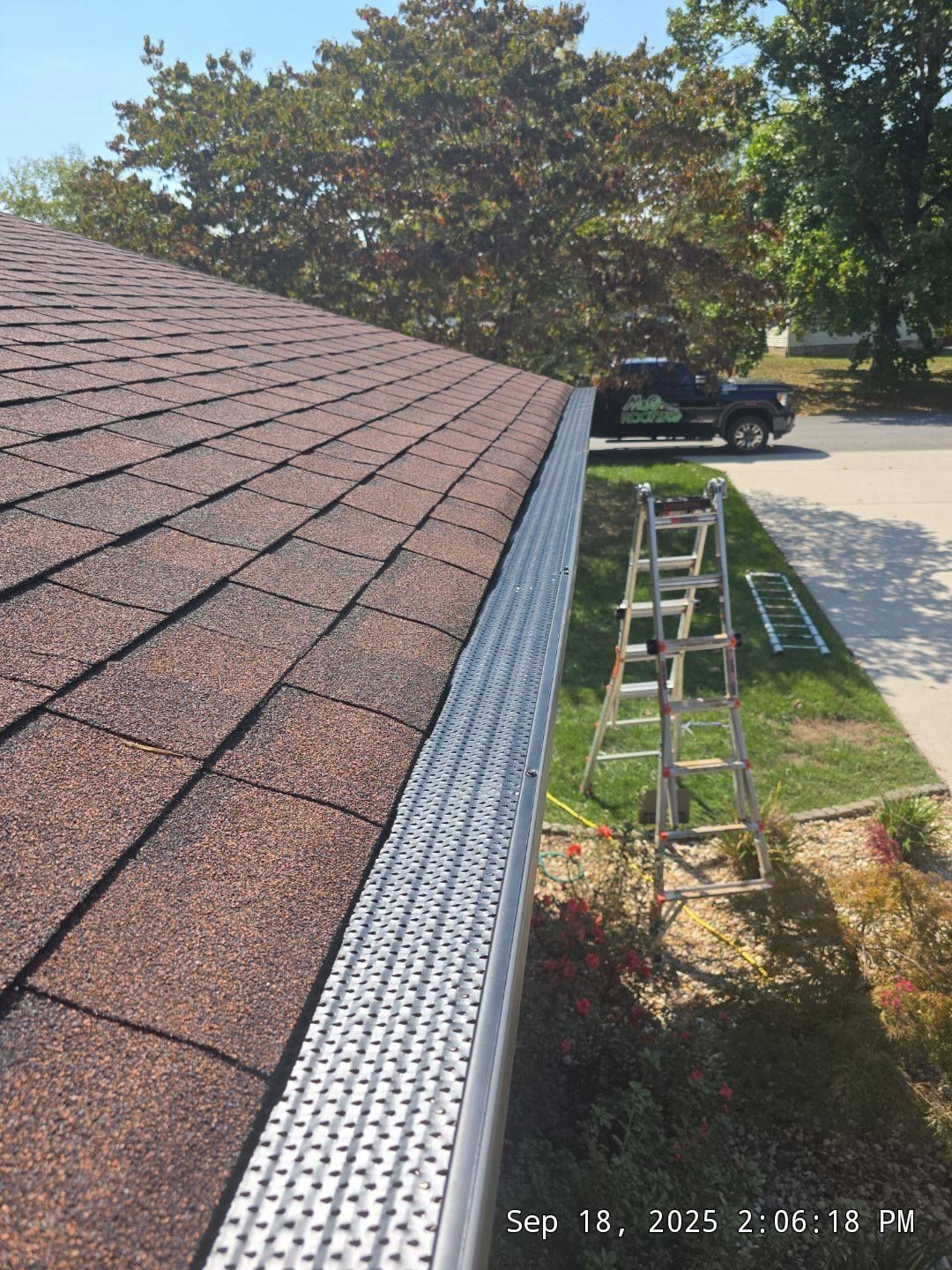 Brown shingled roof with gutter guard, ladder on lawn, black truck in background, sunny day.