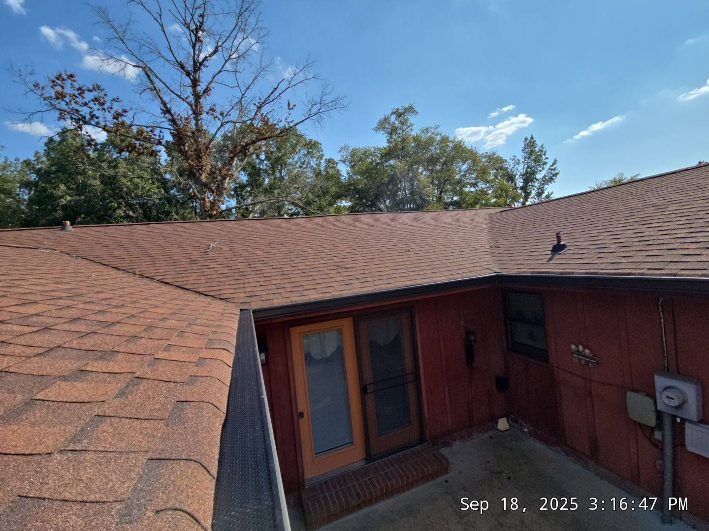 Brown shingled roof with a red building and trees against a blue sky.