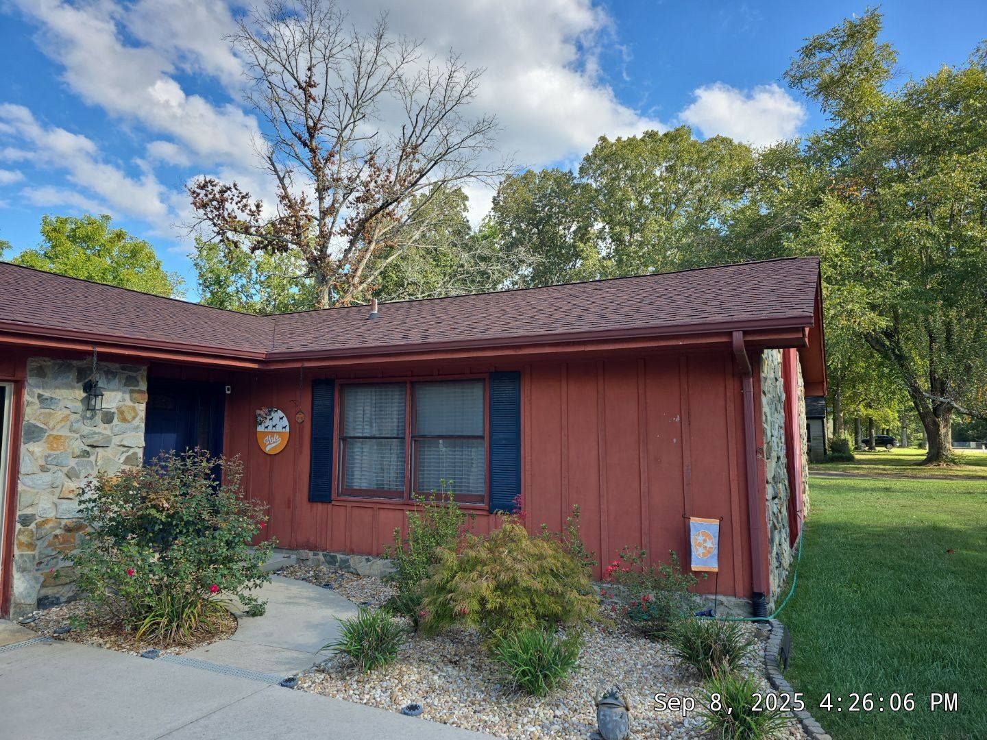 Red-sided house with a red roof, dark blue shutters, stone facade, and landscaping under a blue sky.