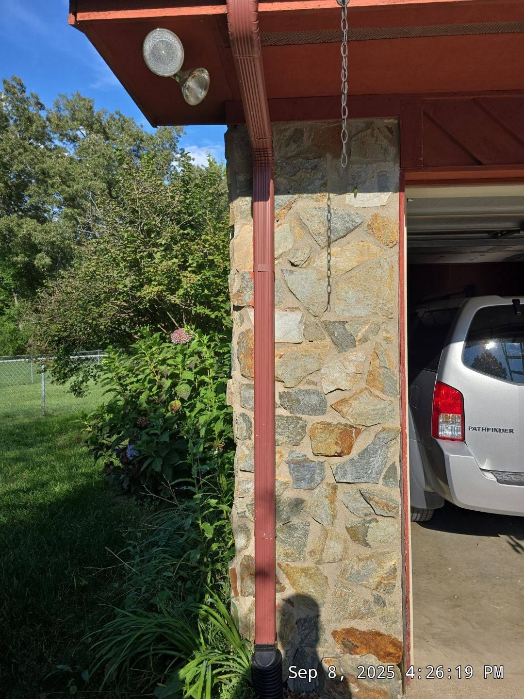 Stone column with brown gutter, chain, and lights. Silver car in garage. Green foliage.