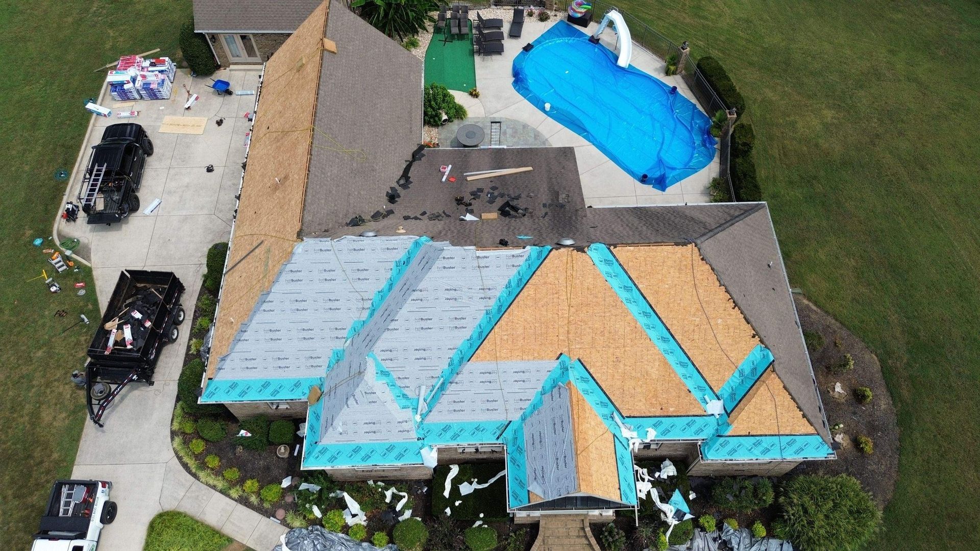 Aerial view of house during roof replacement. Blue tarp covers pool, vehicles parked on driveway.
