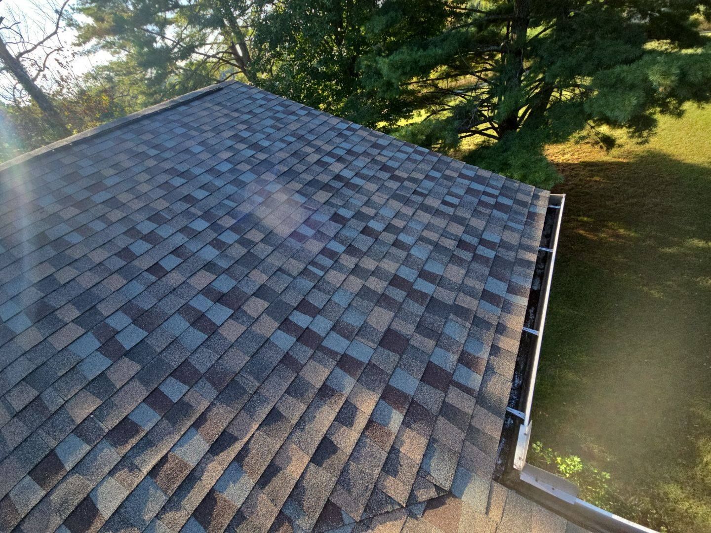 Asphalt shingle roof with blue, brown, and gray hues; edge has a gutter, with trees and grass in the background.